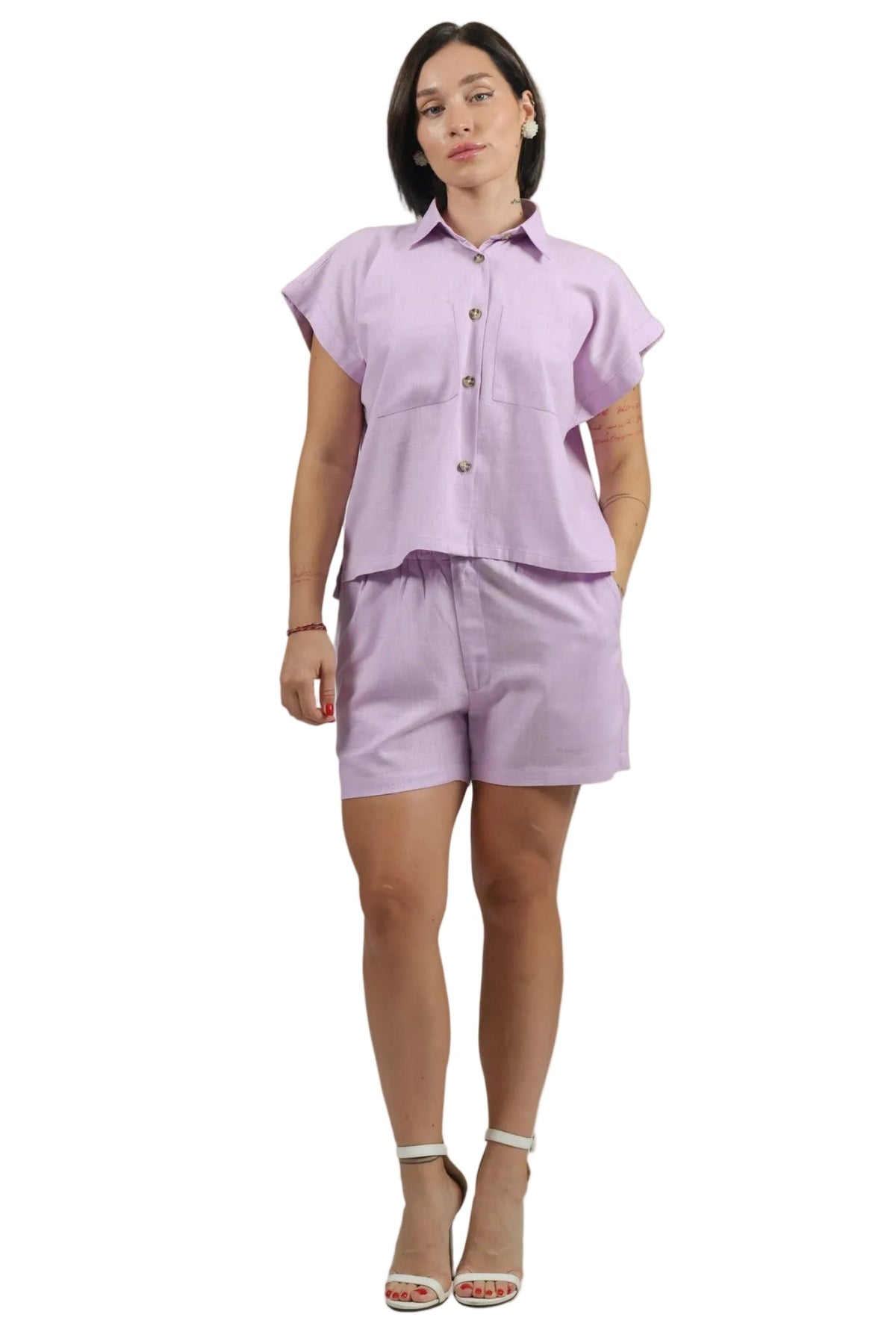A woman modeling a light purple/lavender short-sleeve button-up shirt and matching shorts set, paired with white heeled sandals, against a plain white background.