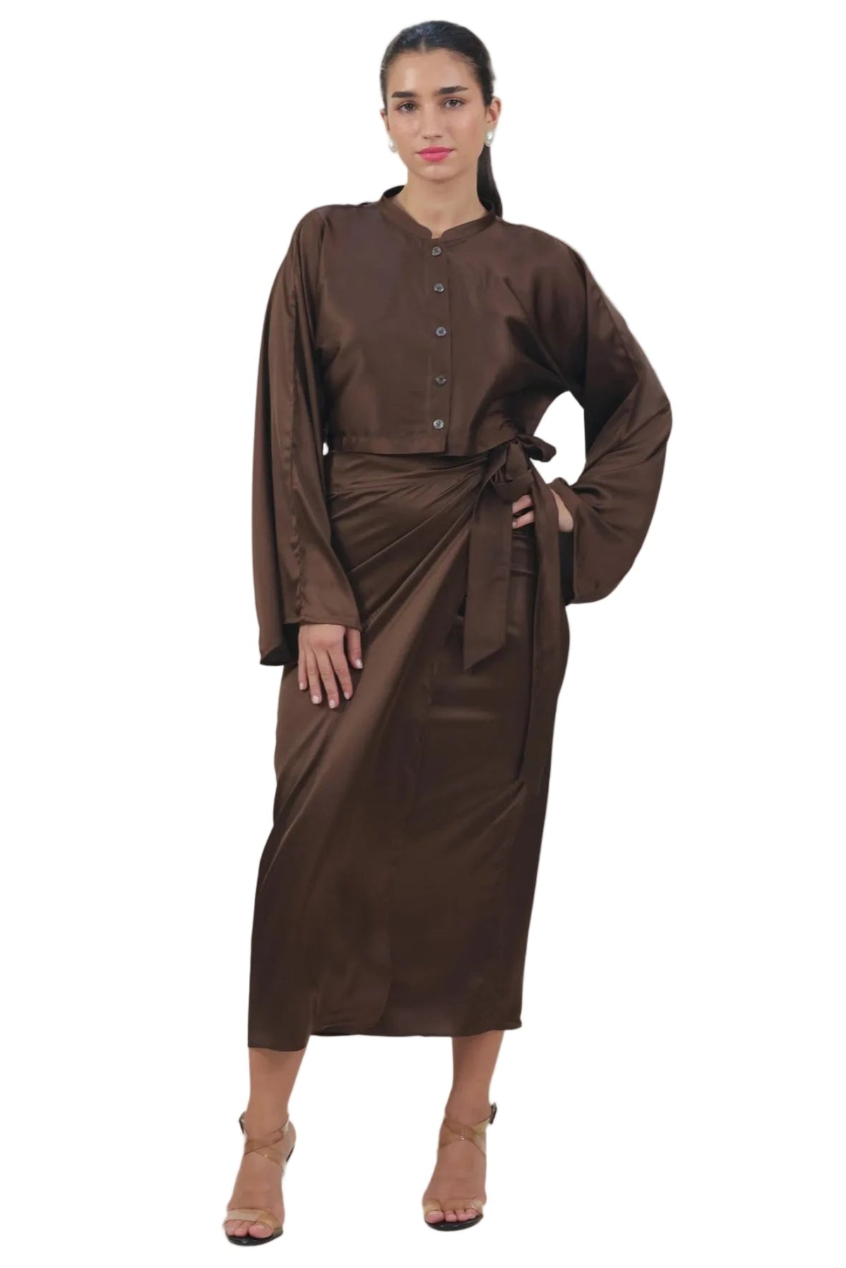 A woman modeling a dark brown long-sleeve dress with a buttoned crop top and a matching draped wrap skirt tied at the waist, photographed against a solid white background.