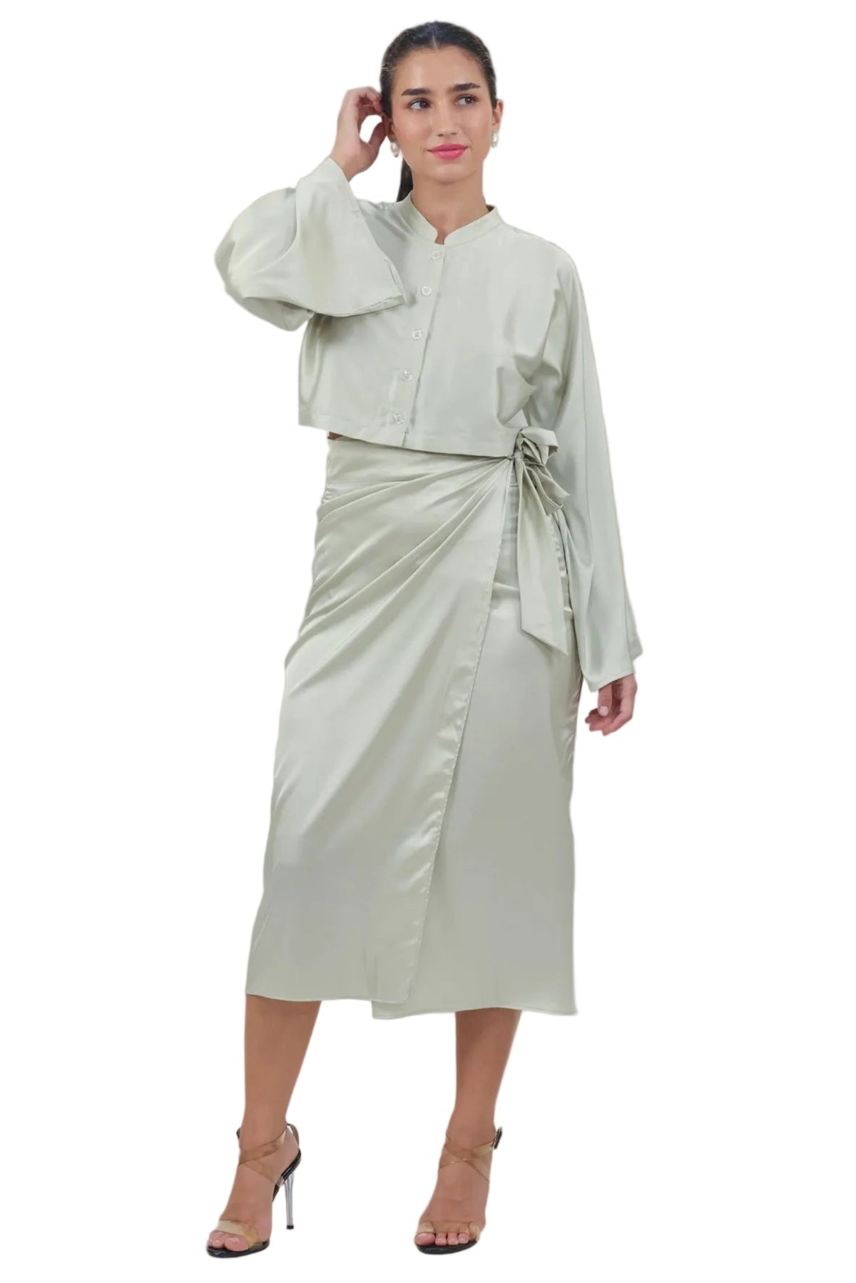 A woman modeling a light sage green two-piece outfit consisting of a long-sleeve cropped jacket with a stand collar and a matching wrap midi skirt with a tie waist, paired with nude high-heeled sandals against a plain white background.