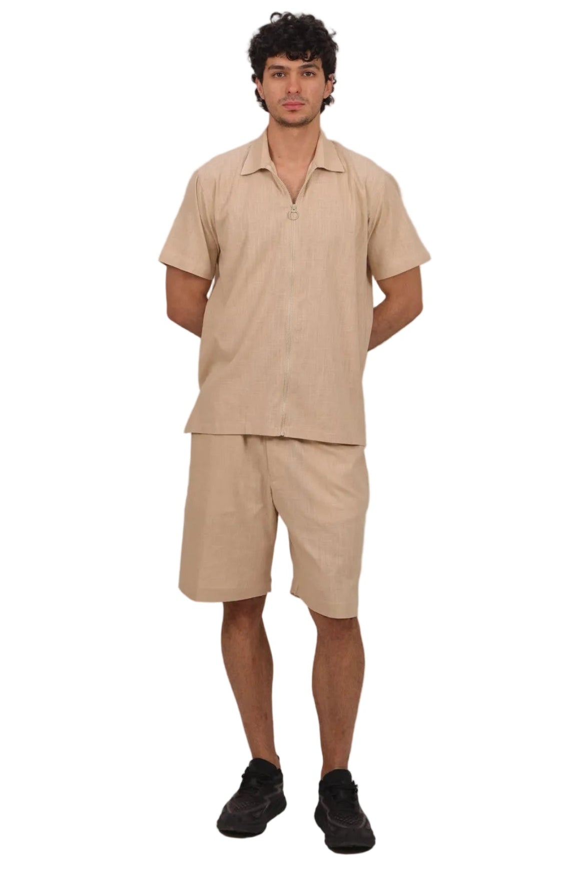 A man modeling a matching beige short-sleeve button-up shirt and shorts set, paired with black sneakers, against a solid white background.