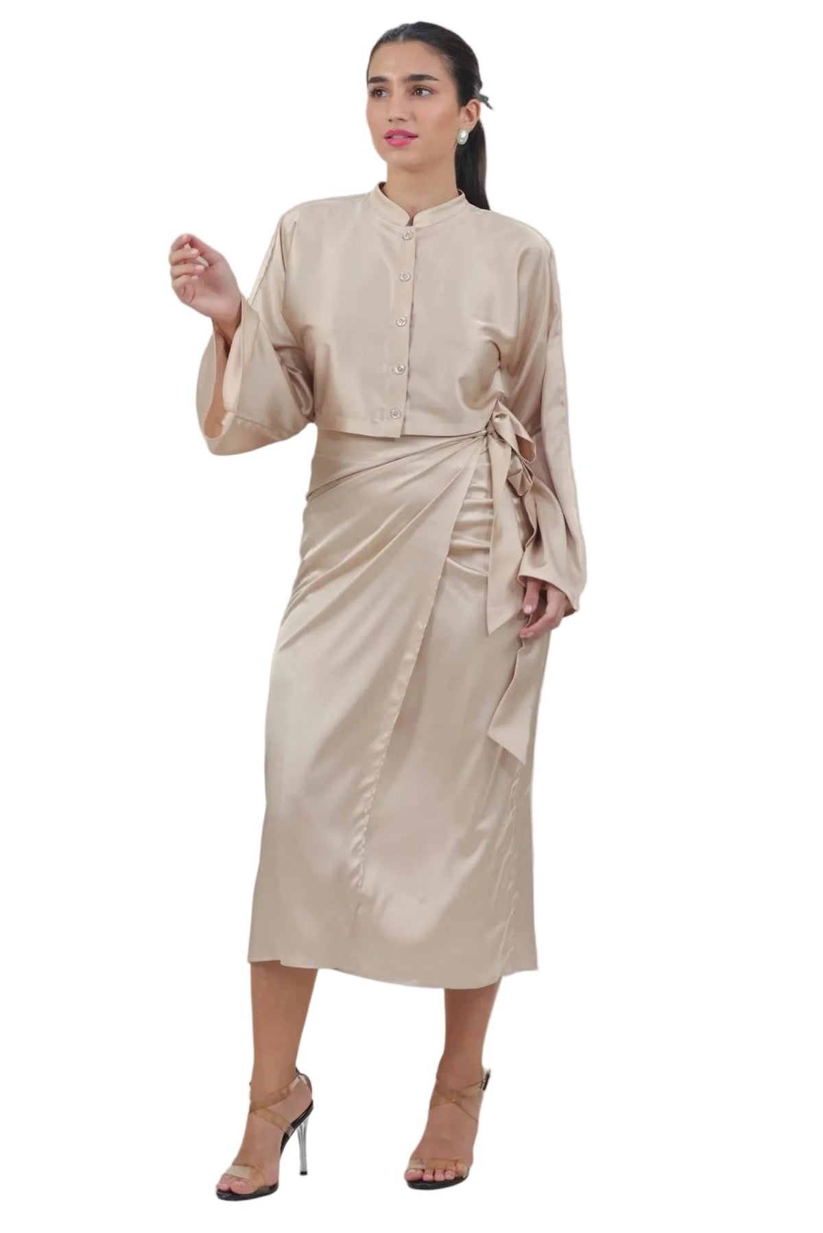 A woman modeling a beige satin midi dress with a high collar, long sleeves, and a side tie, paired with beige high-heeled sandals, set against a plain white background.