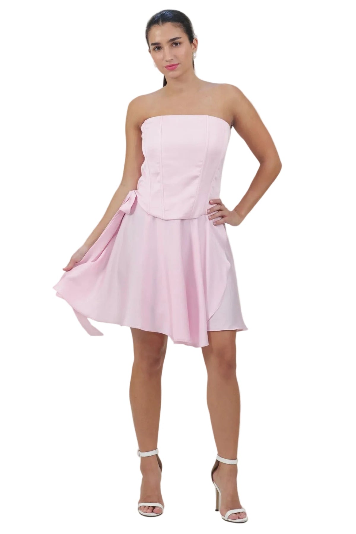 A woman modeling a light pink strapless mini dress with a fitted bodice and flared skirt, paired with white high-heeled sandals, against a plain white background.