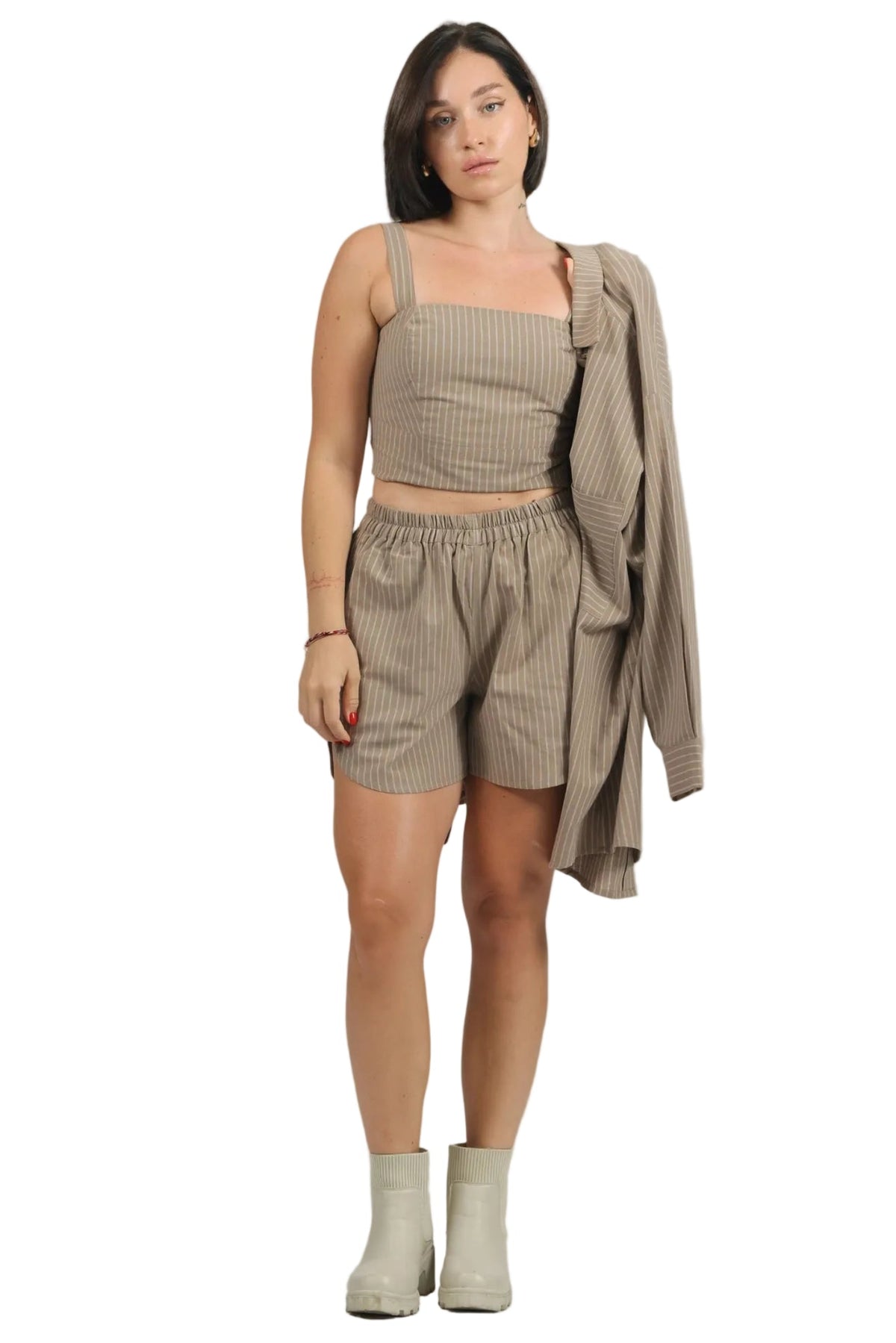 A woman modeling a matching beige and white vertically striped two-piece set consisting of a cropped tank top and shorts, with a matching long-sleeve shirt draped over her shoulder, set against a plain white background.