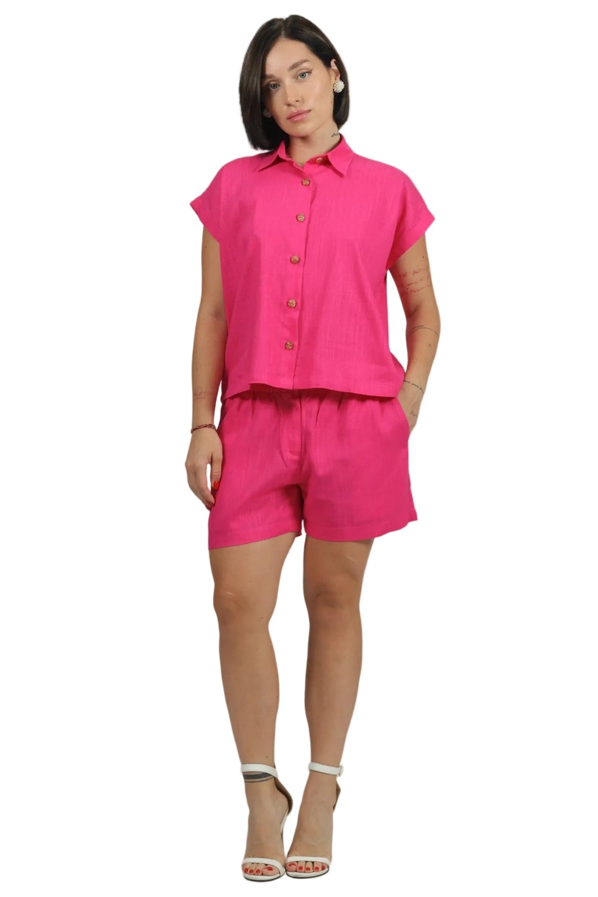 A woman modeling a bright pink two-piece set consisting of a short-sleeve button-up shirt and matching shorts, paired with white heeled sandals, against a plain white background.
