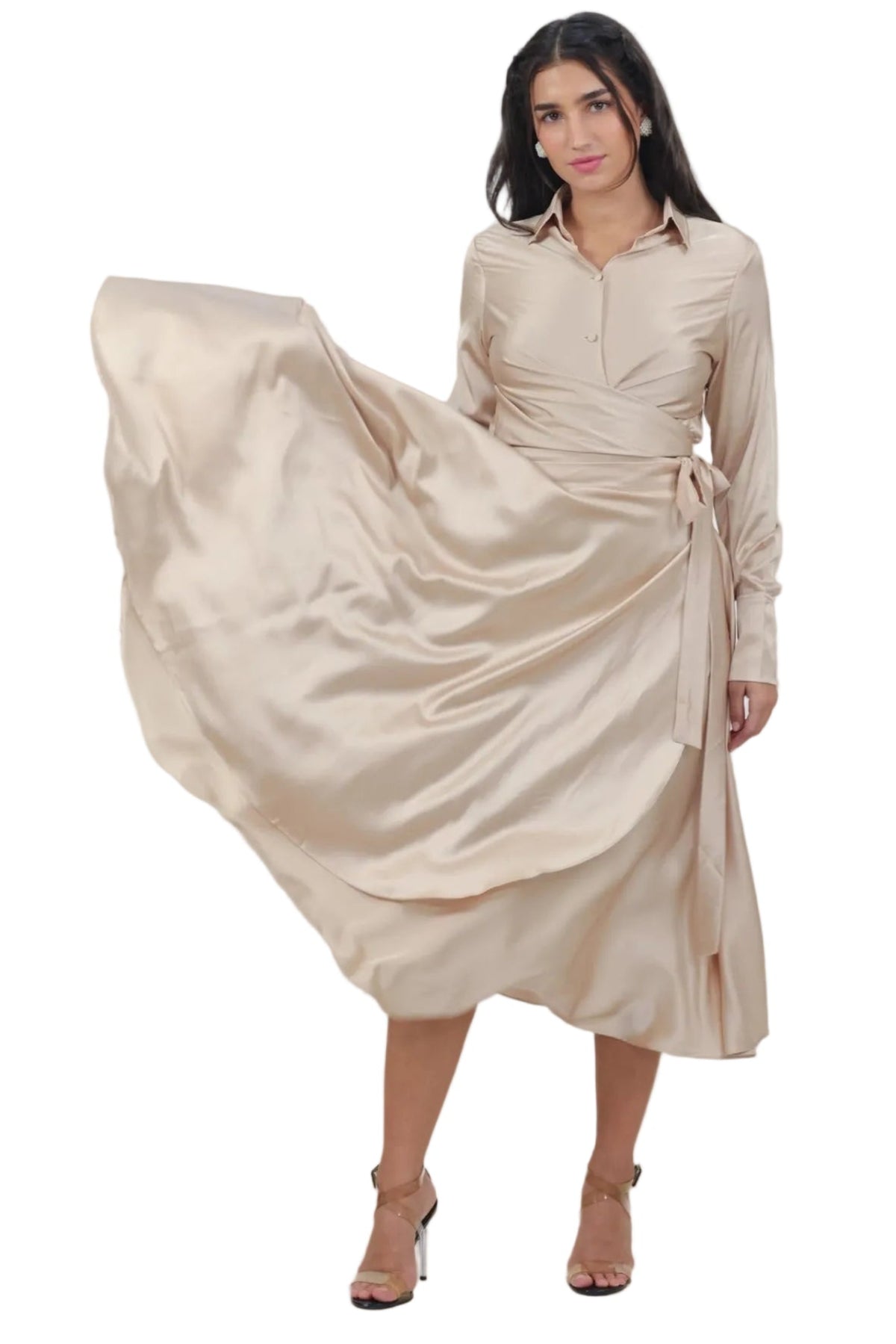 A woman modeling a long-sleeve beige satin midi dress with a wrap-style waist and tie belt, captured mid-motion as she swirls the fabric. The background is a solid, warm neutral beige studio backdrop.