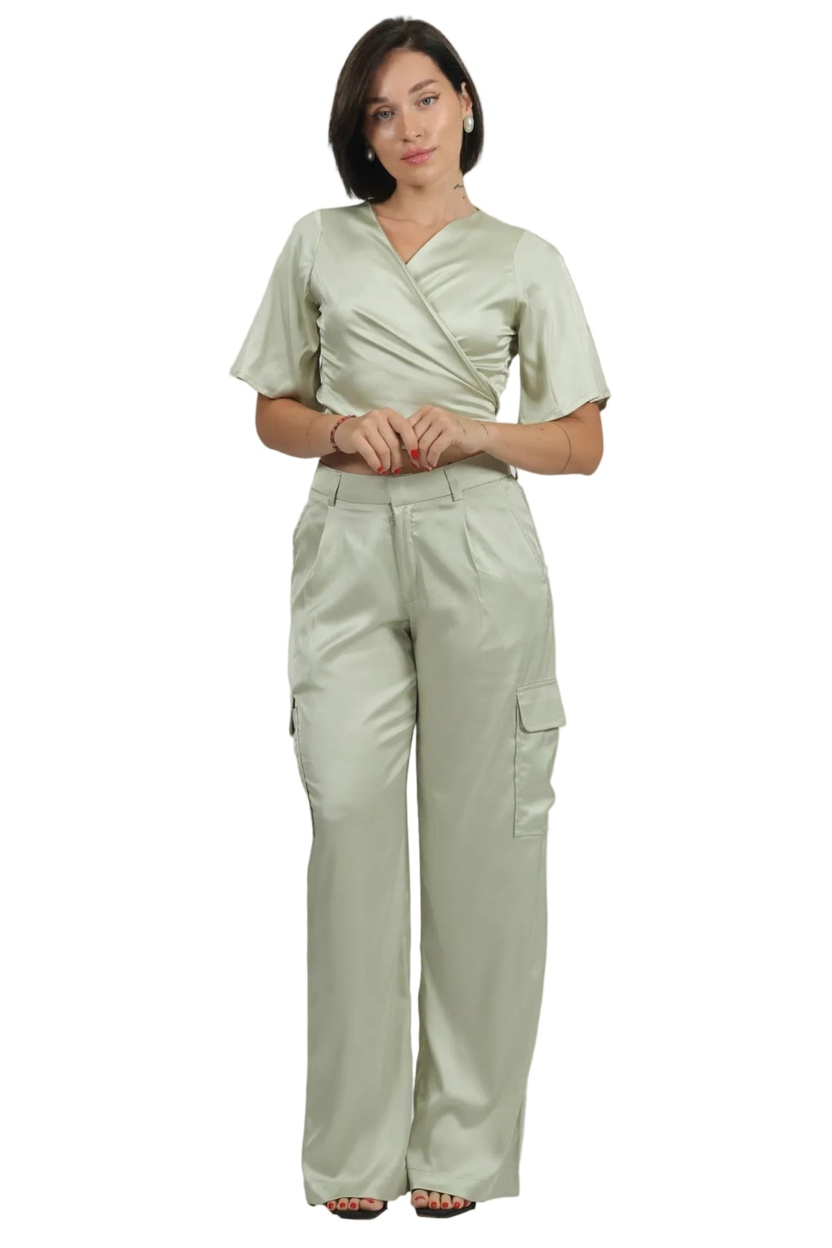 A woman modeling a light sage green scrub set consisting of a short-sleeve wrap-style top and matching high-waisted cargo pants, shot against a plain white background.