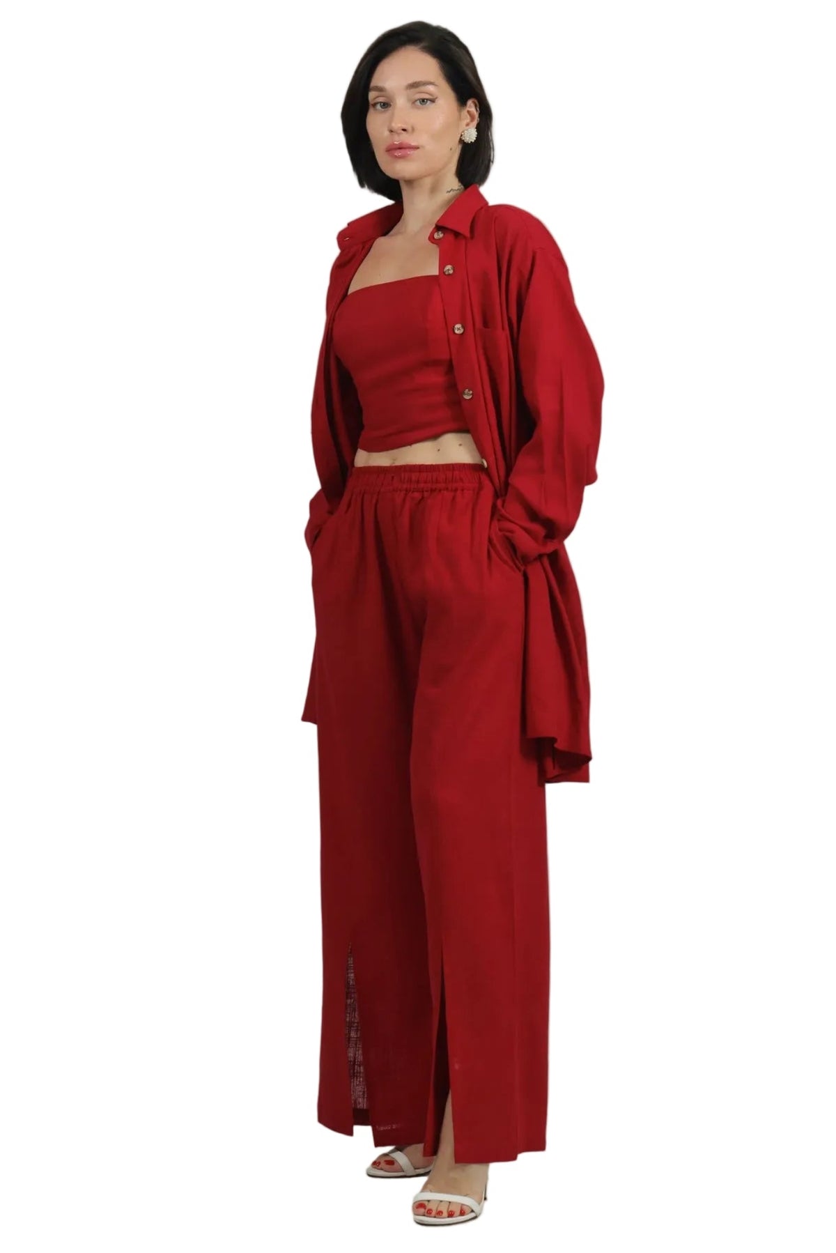 A woman modeling a matching red two-piece outfit consisting of a cropped tank top and wide-leg trousers, styled with an open button-up shirt and white heeled sandals against a plain white background.