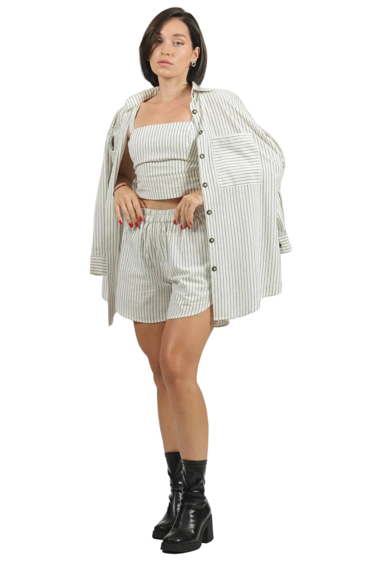 A woman modeling a matching two-piece set consisting of a striped crop top and shorts, worn under an oversized matching striped button-up shirt, paired with black ankle boots, on a solid white background.