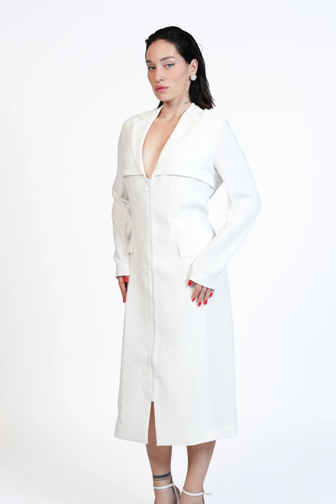 A woman modeling a white long-sleeve midi dress with a deep V-neckline and front slit, paired with white strappy high-heeled sandals, set against a plain white studio background.