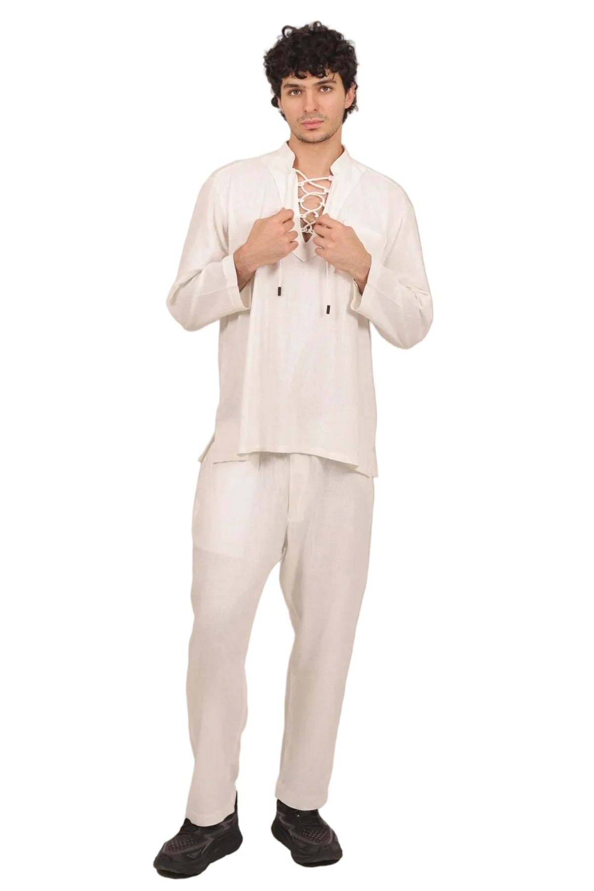 A man modeling a cream-colored two-piece traditional outfit consisting of a long-sleeve top with lace-up front details and matching trousers, set against a solid white background.