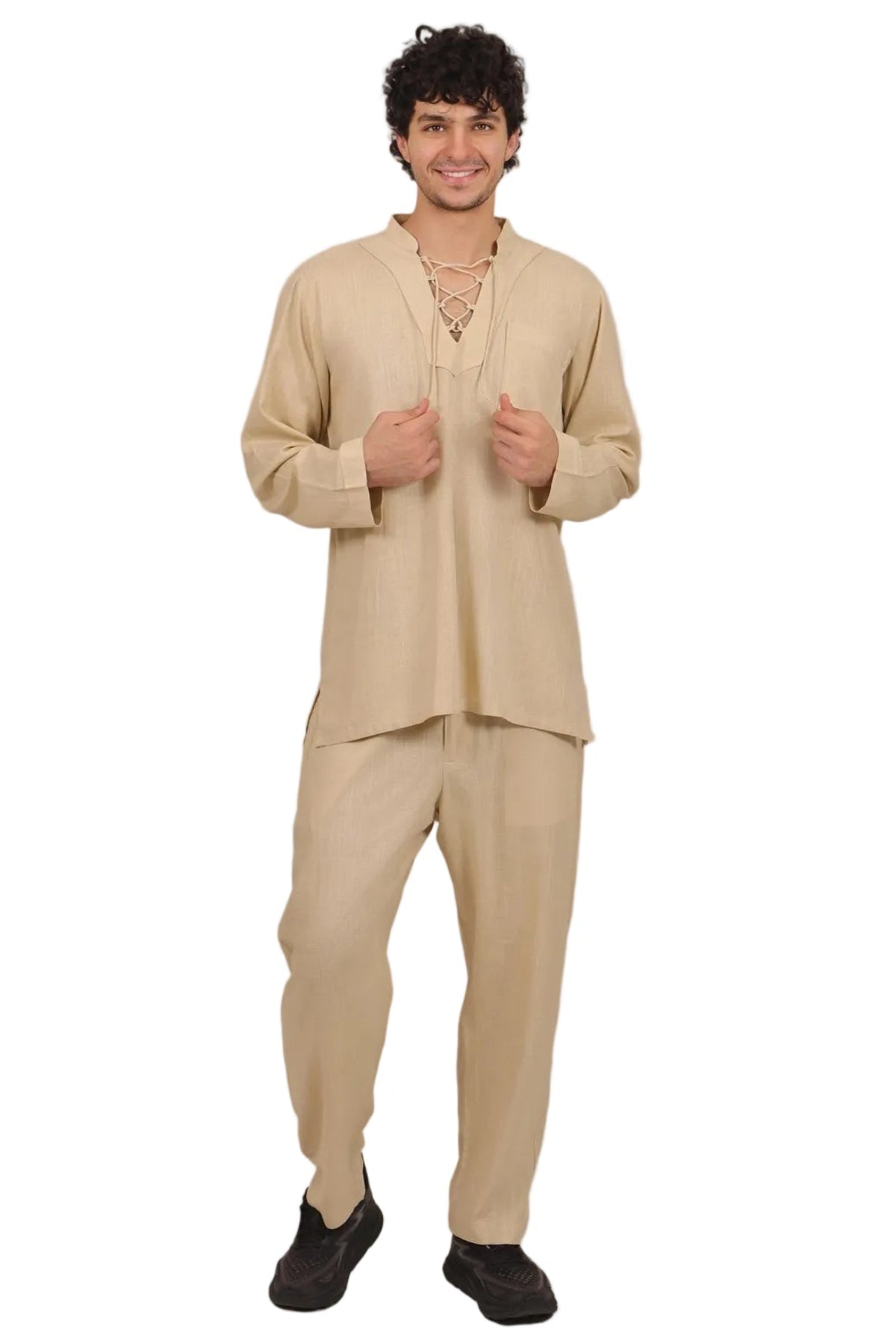 A man modeling a beige two-piece traditional outfit consisting of a long-sleeve tunic with a split neckline and matching loose pants, paired with black shoes, on a solid white background.