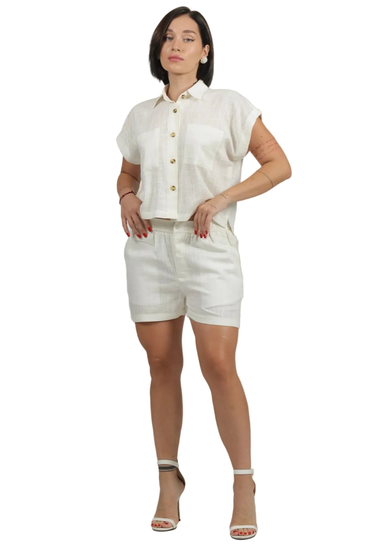 A woman modeling a matching off-white linen two-piece set consisting of a short-sleeve button-up shirt and high-waisted shorts, paired with beige high-heeled sandals against a solid white background.