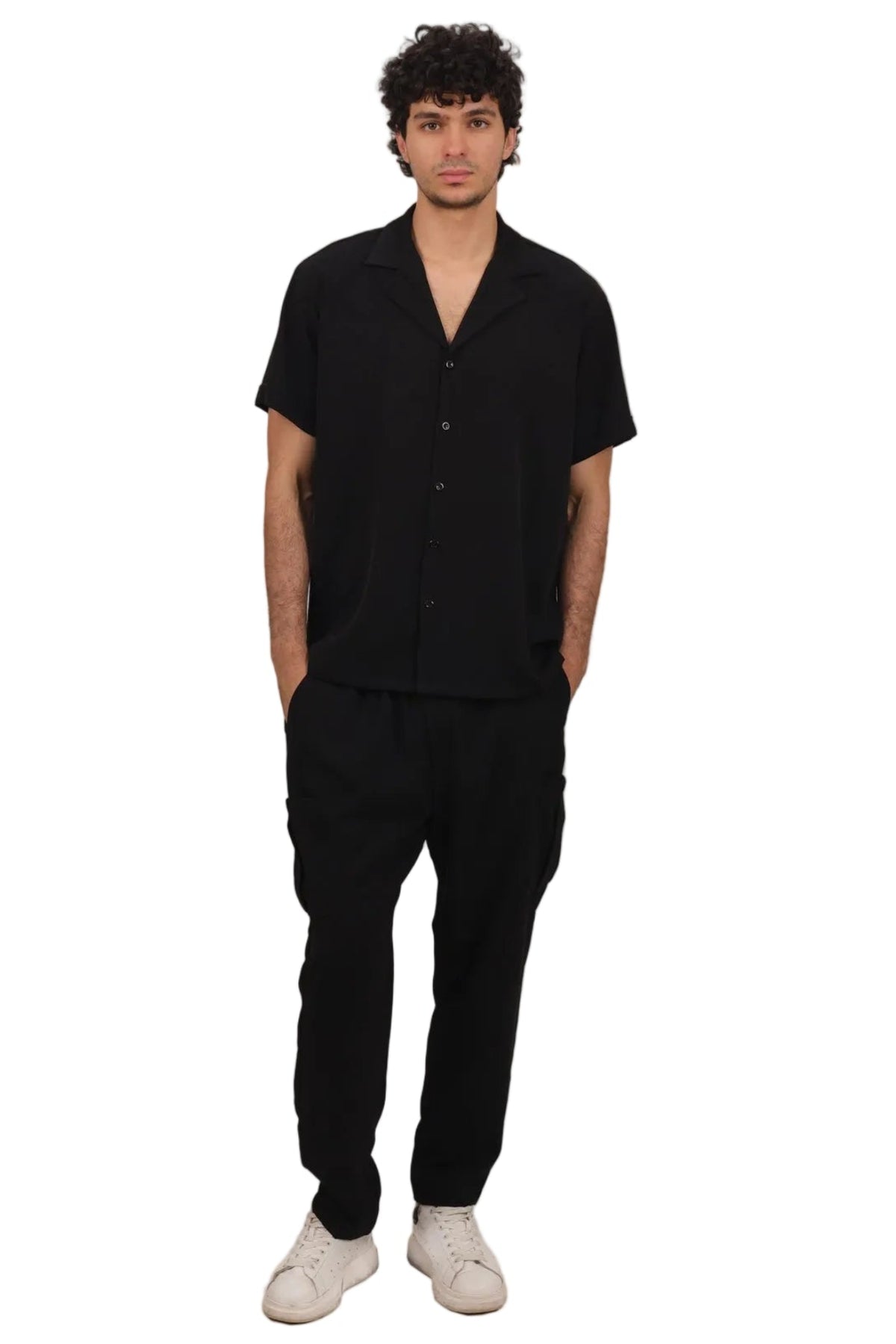 A man modeling a black short-sleeve button-up shirt and black trousers with white sneakers, standing against a solid white background.