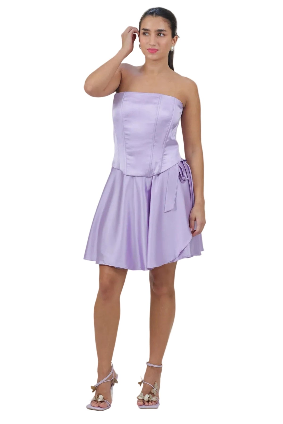 A woman modeling a lavender strapless taffeta mini dress with a corset-style bodice and flared skirt, paired with metallic high-heeled sandals, against a plain white background.