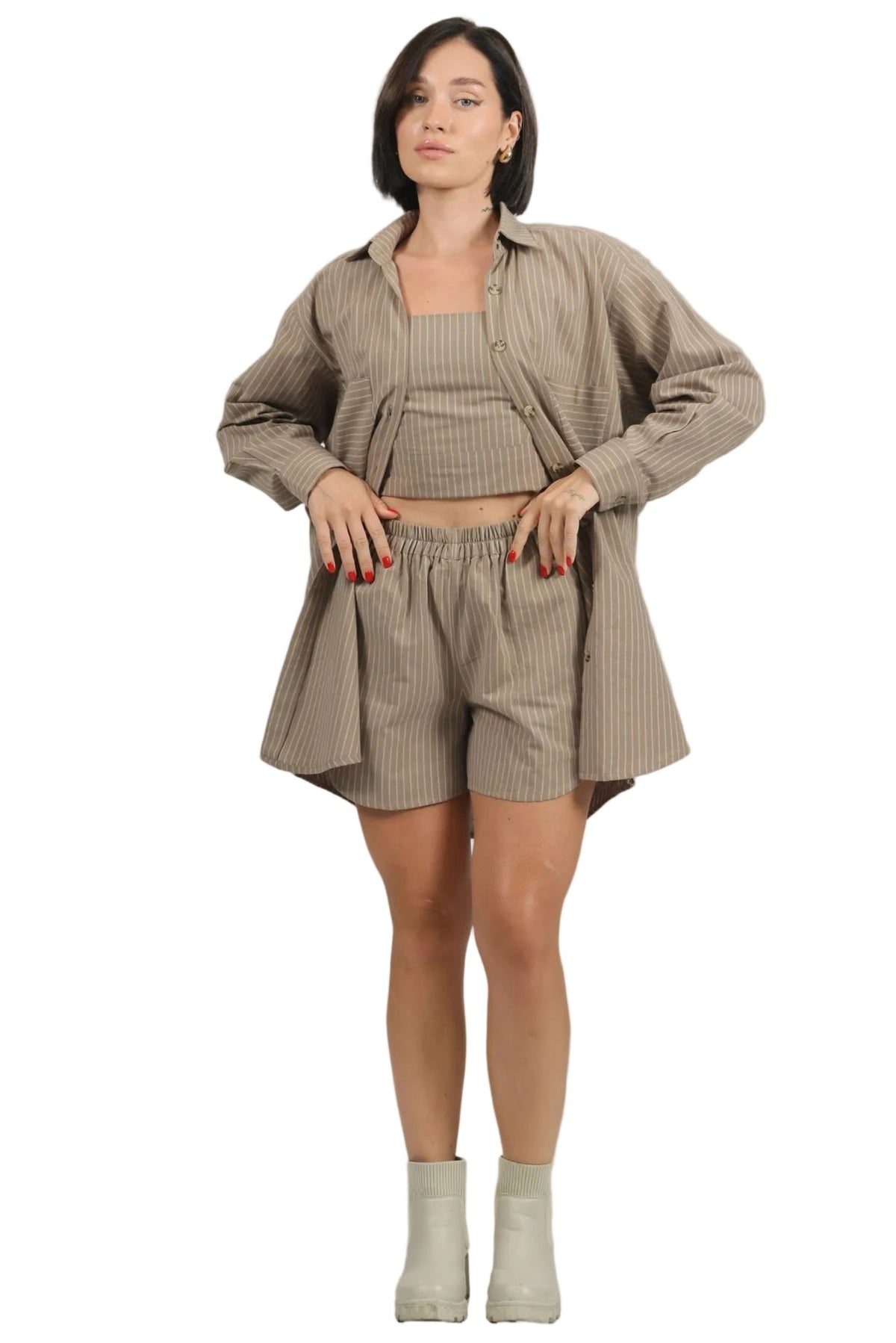 A woman modeling a matching beige pinstriped lounge set consisting of a cropped tank top, shorts, and an oversized button-up shirt, paired with light beige ankle boots against a plain white background.