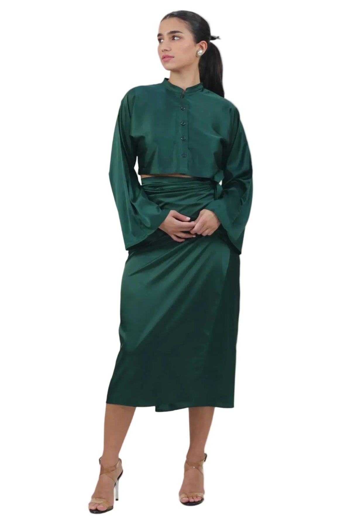 A woman modeling a dark green satin two-piece set consisting of a long-sleeve button-up top and a matching midi skirt, paired with beige high-heeled sandals against a plain white background.