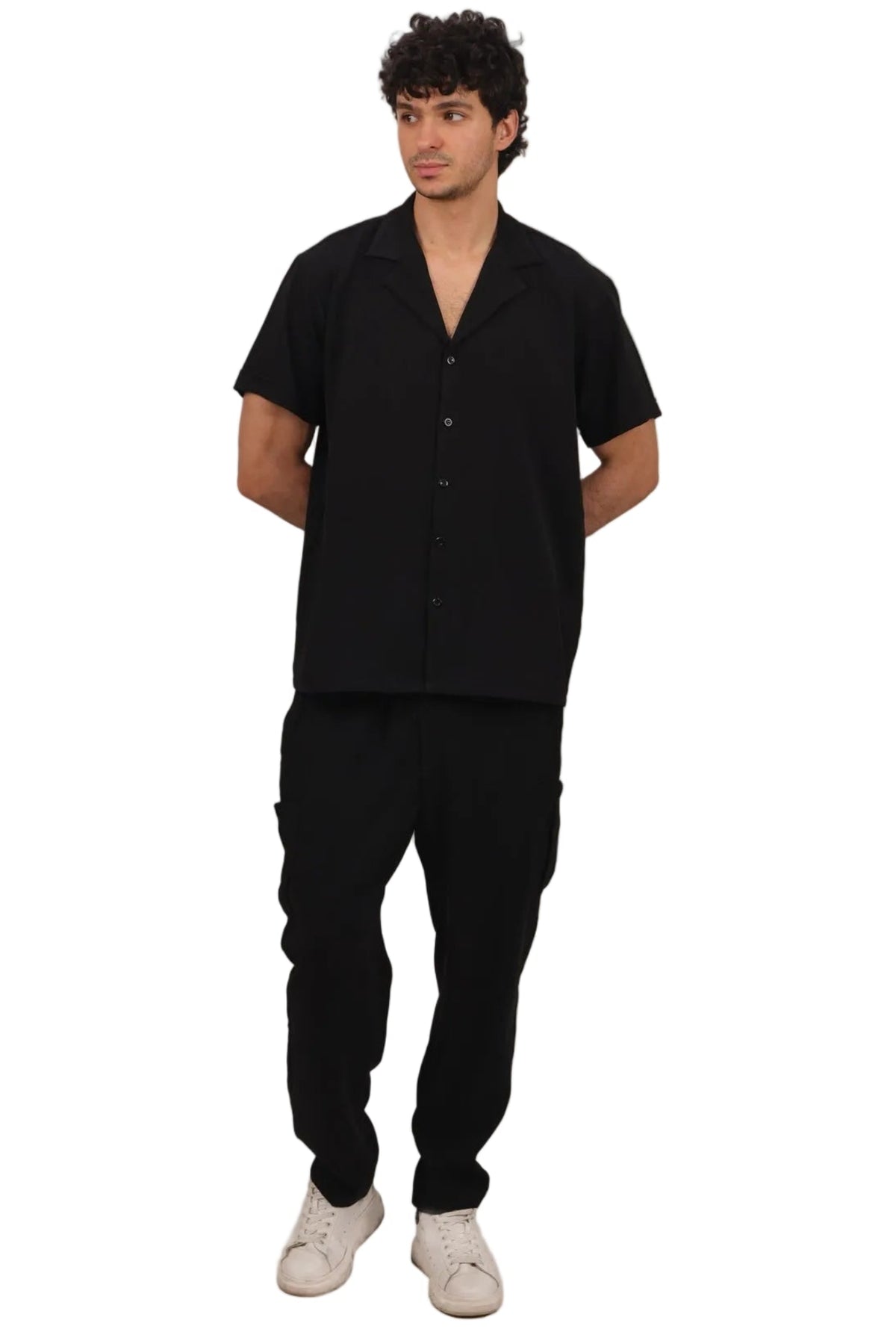 A man modeling a black short-sleeve button-up shirt and matching black pants, paired with white sneakers, against a solid white background.