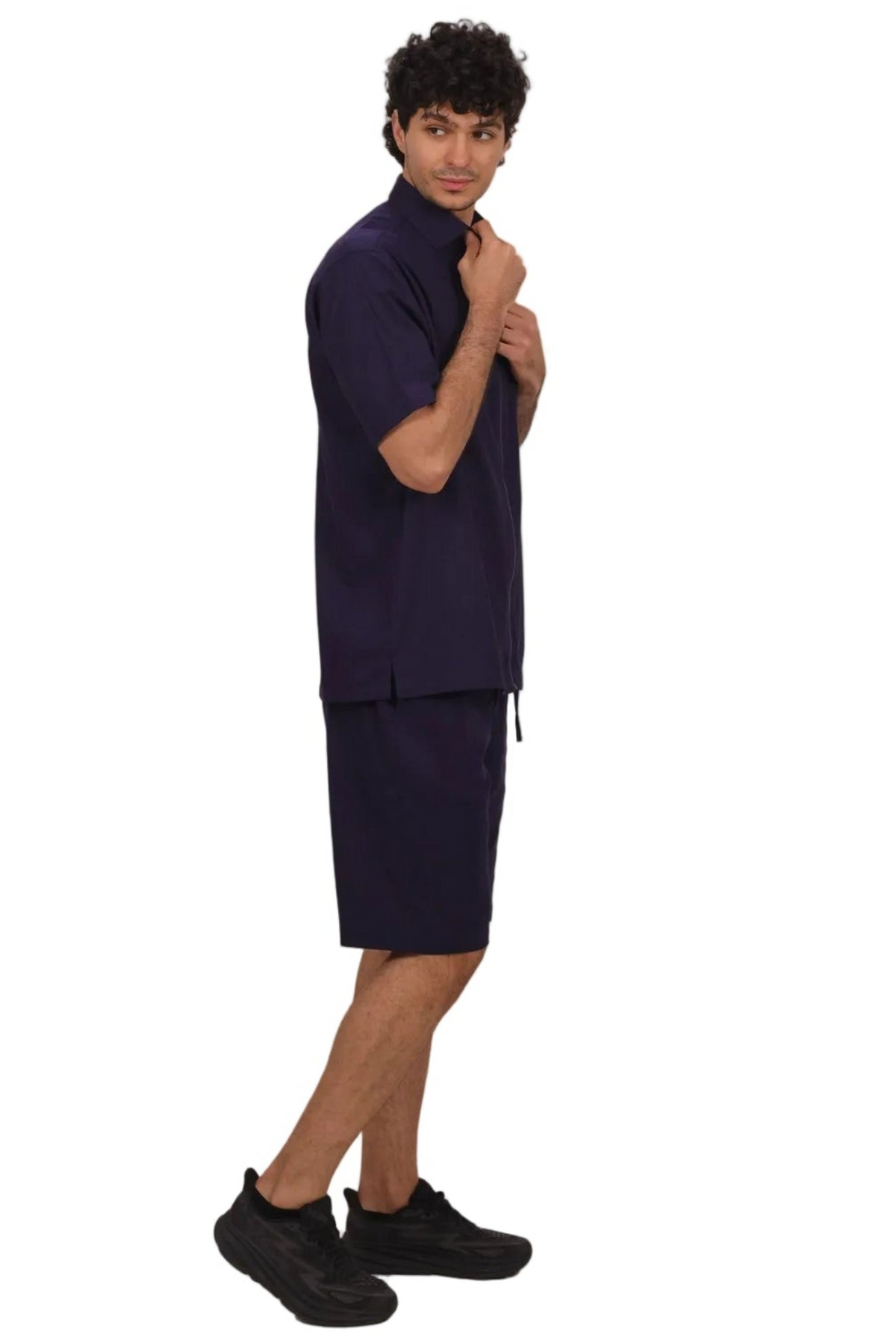 A man modeling a dark navy blue two-piece lounge set consisting of a short-sleeve button-up shirt and matching shorts, paired with black sneakers, against a solid white background.