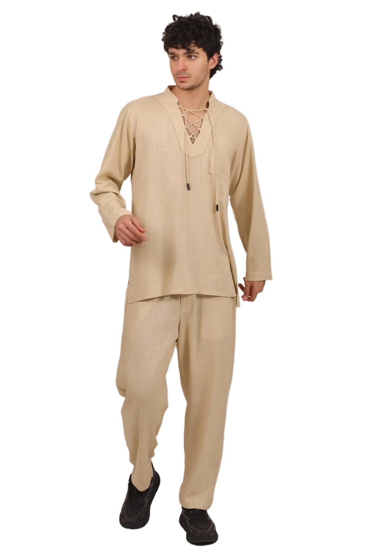 A man modeling a beige two-piece traditional outfit consisting of a long-sleeve tunic with a lace-up neckline and matching loose trousers, paired with black shoes, against a solid white background.