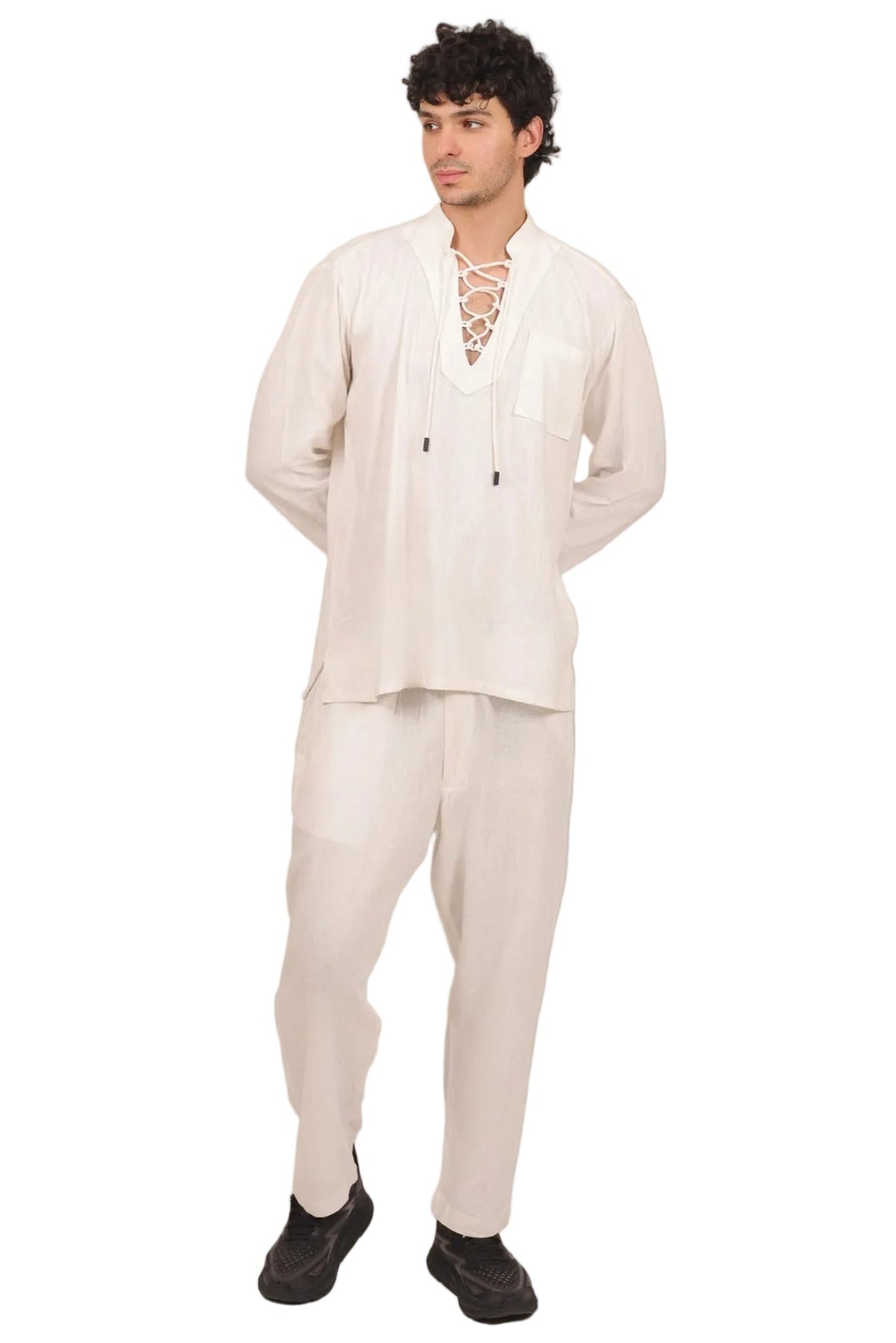 A man modeling a cream-colored two-piece traditional outfit consisting of a long-sleeve tunic with a lace-up neckline and matching trousers, paired with black shoes, against a solid white background.
