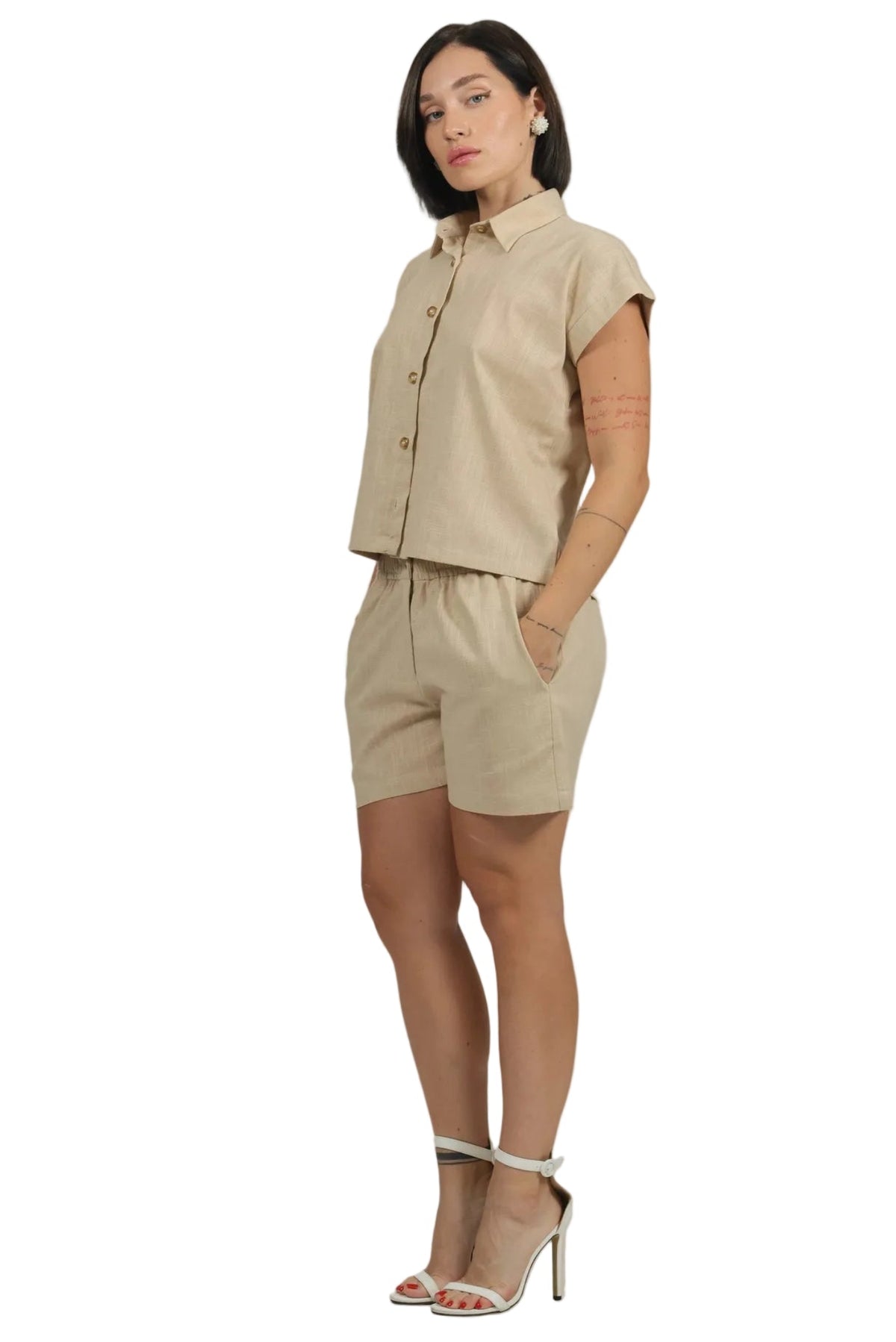 A woman modeling a light beige linen two-piece set consisting of a short-sleeve button-up shirt and matching shorts, paired with white high-heeled sandals, against a solid white background.