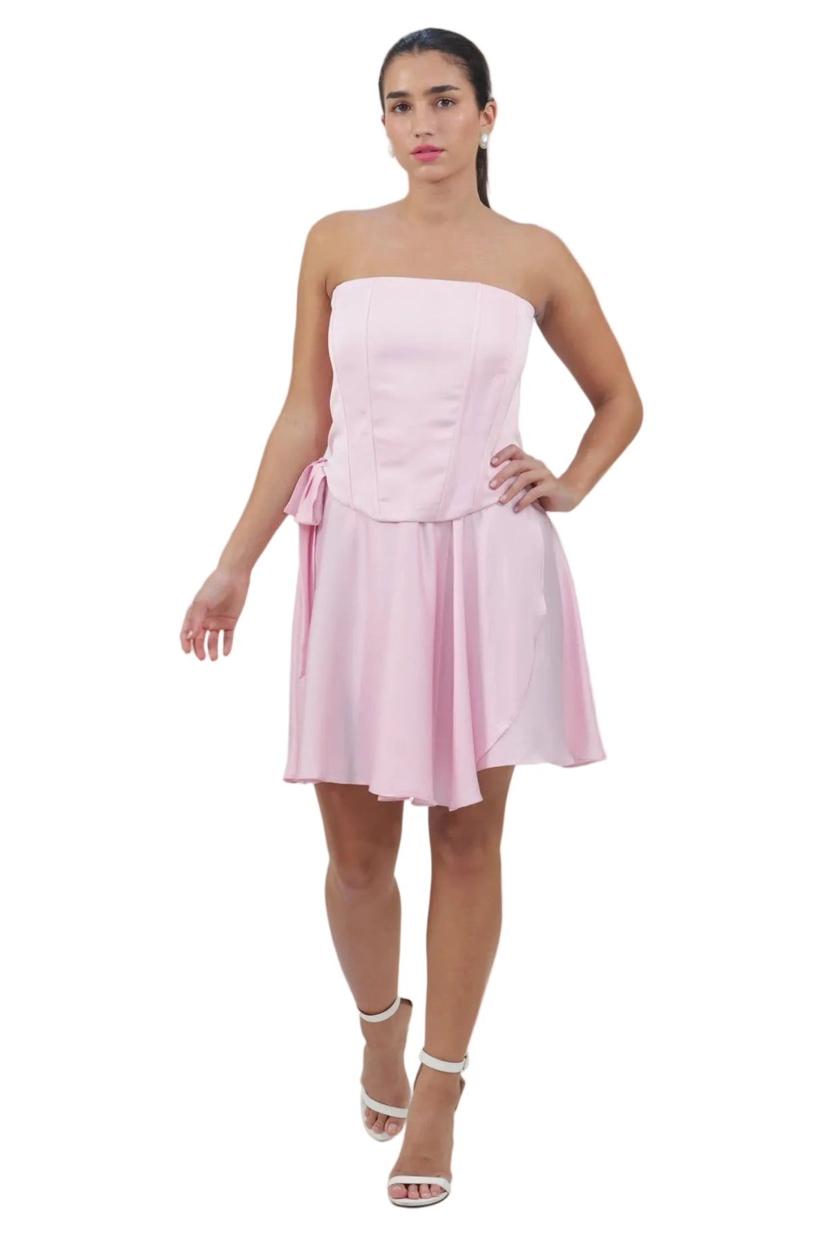 A woman modeling a light pink strapless mini dress with a structured corset-style bodice and a flared skirt, paired with white heeled sandals, against a plain white background.