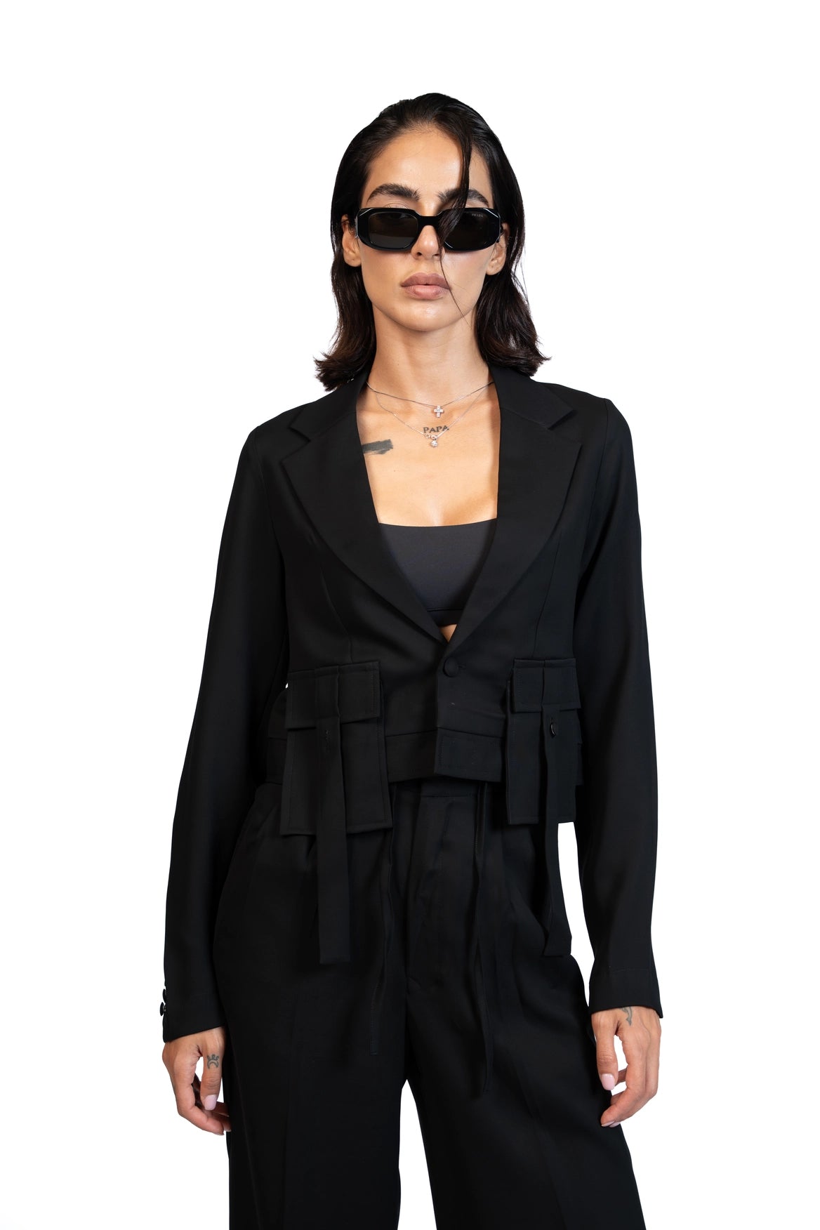 A woman modeling a black tailored blazer with structured shoulders and draped tie details at the waist, paired with matching black trousers and black sunglasses, against a plain white background.