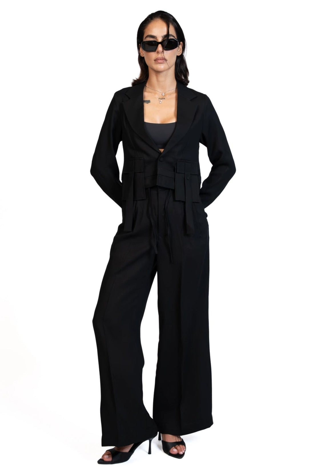 A model wearing a black tailored jumpsuit with a draped waist, wide legs, and black pointed-toe heels, photographed against a plain white background.