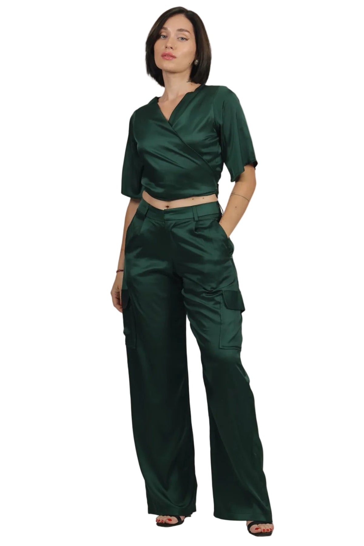 A woman modeling a dark green satin two-piece set consisting of a short-sleeve wrap-style crop top and matching wide-leg cargo pants, set against a plain white background.