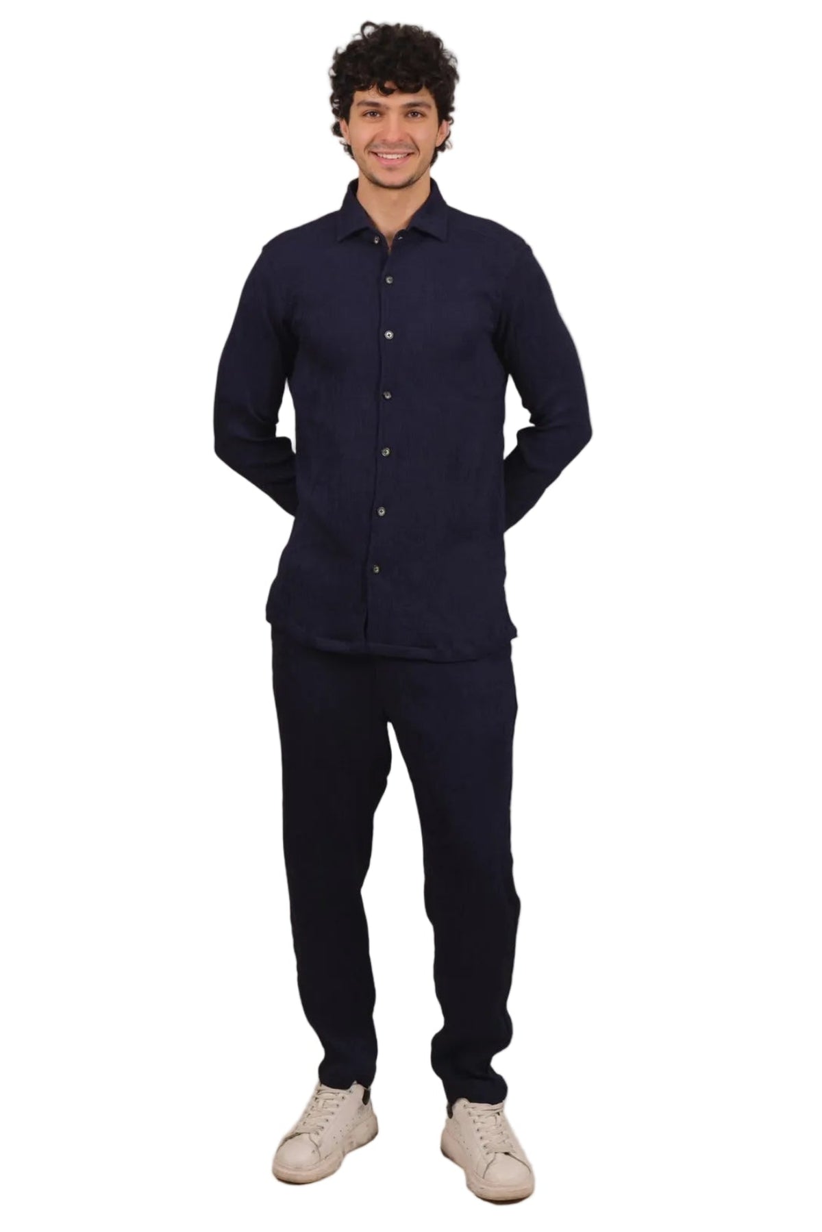 A man wearing a dark navy blue long-sleeve button-up shirt and matching dark pants, paired with off-white sneakers, standing against a solid white background.