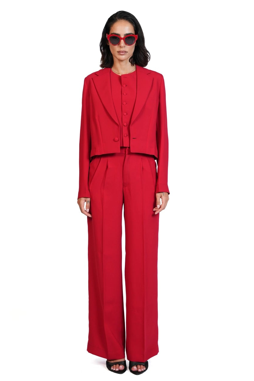 A model wearing a bright red tailored suit consisting of a long-sleeve blazer with a unique draped neckline and matching wide-leg trousers, styled with red-tinted sunglasses and black sandals against a plain white background.