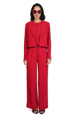 A model wearing a bright red tailored suit consisting of a long-sleeve blazer with a unique draped neckline and matching wide-leg trousers, styled with red-tinted sunglasses and black sandals against a plain white background.