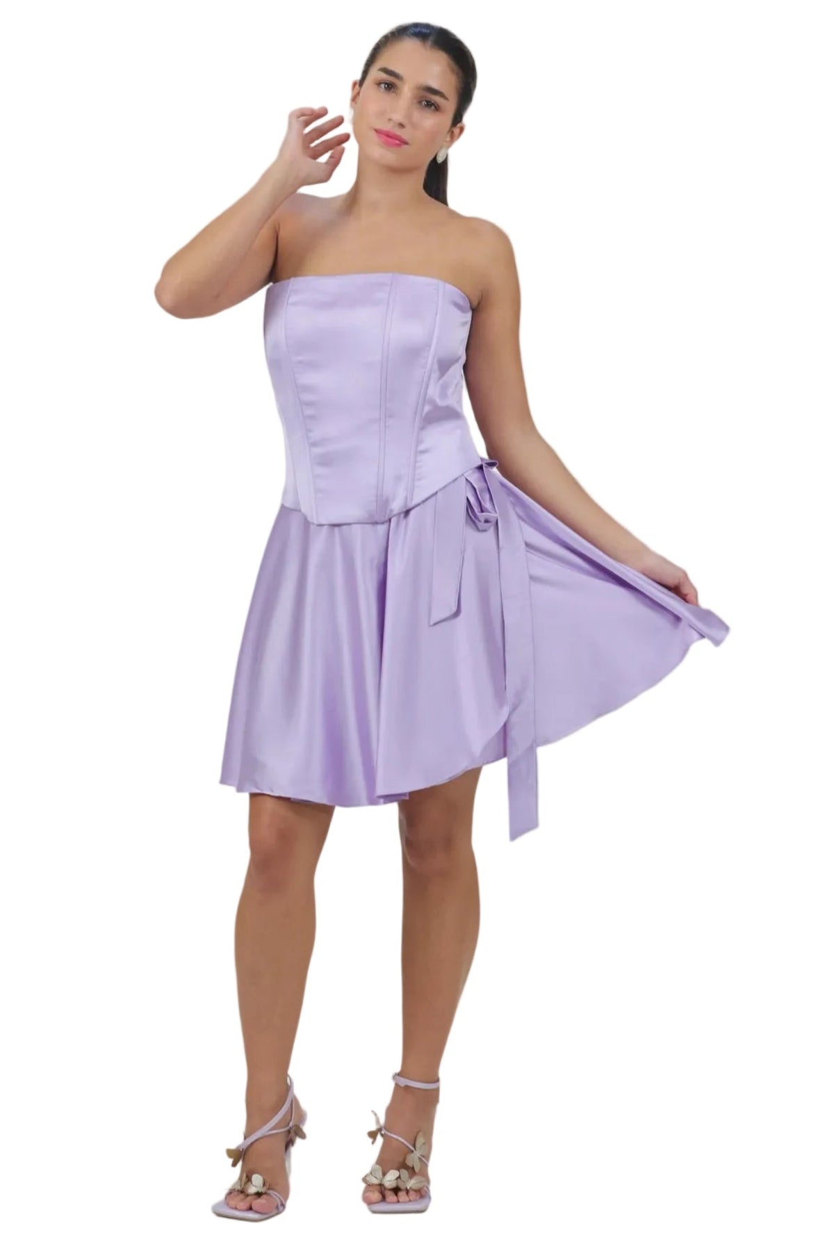 A woman modeling a lavender strapless mini dress with a structured corset-style bodice and a flared skirt, holding out one side of the skirt to show its drape against a plain white background.
