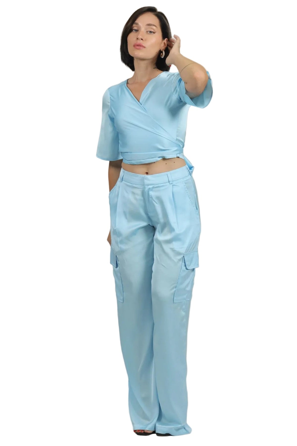A woman modeling a light blue two-piece scrub set consisting of a short-sleeve wrap-style crop top and matching wide-leg cargo pants, set against a plain white background.