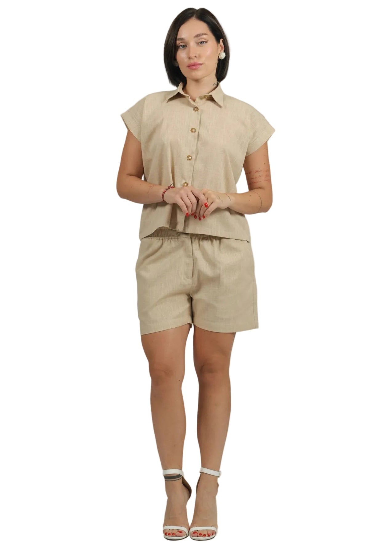 A woman modeling a light beige linen two-piece set consisting of a short-sleeve button-up shirt and matching shorts, paired with beige high-heeled sandals, against a solid white background.
