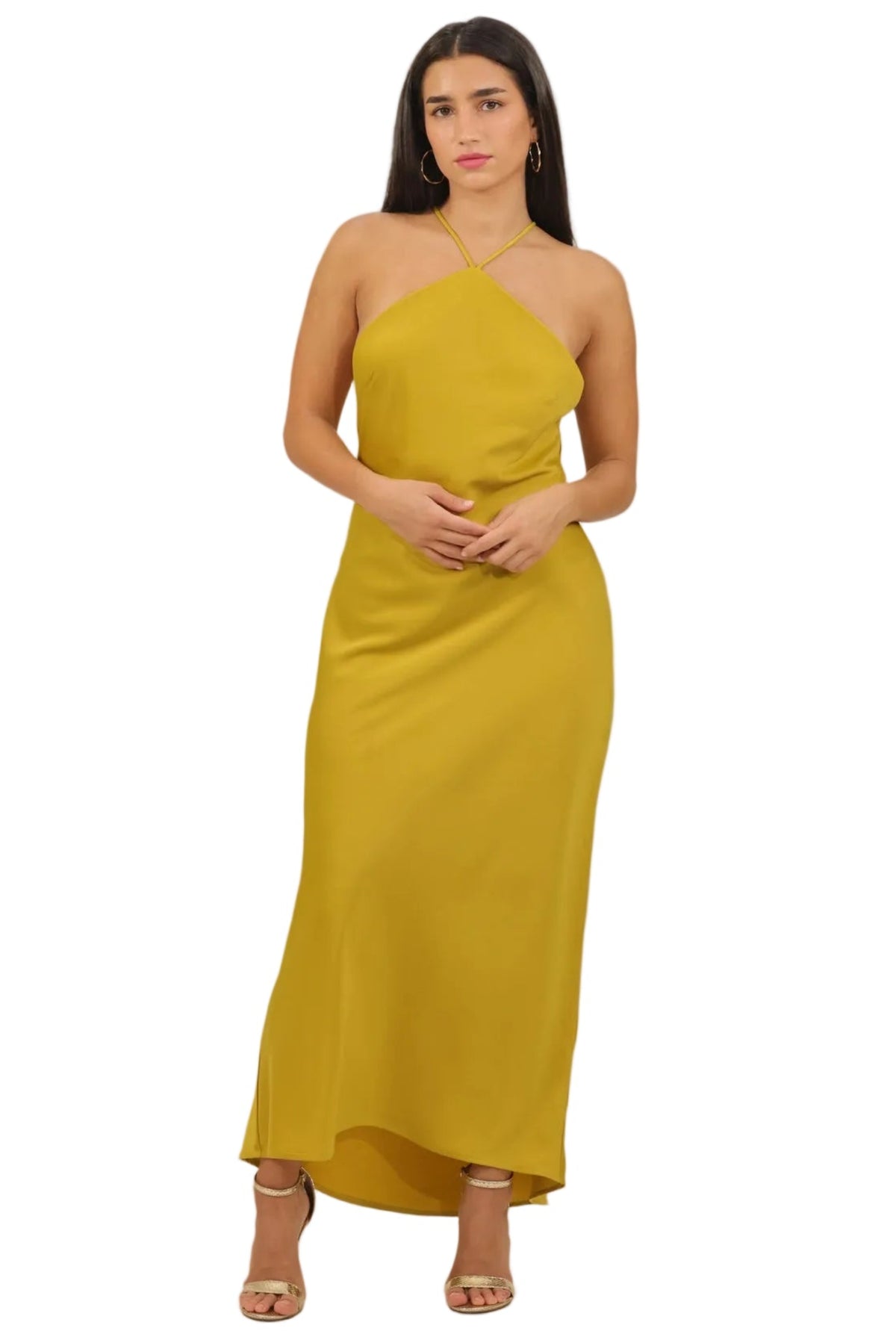 A woman modeling a mustard yellow halter-neck maxi dress with a fitted silhouette and a slight flare at the hem, paired with clear high-heeled sandals, set against a plain white background.