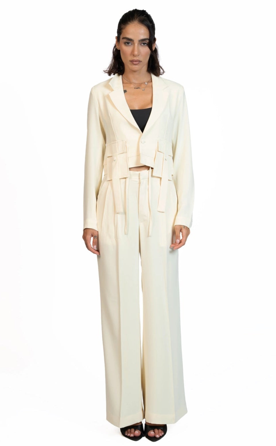 A woman stands against a plain white studio background wearing a cream-colored tailored jumpsuit with a belted waist and wide legs, paired with dark sandals.