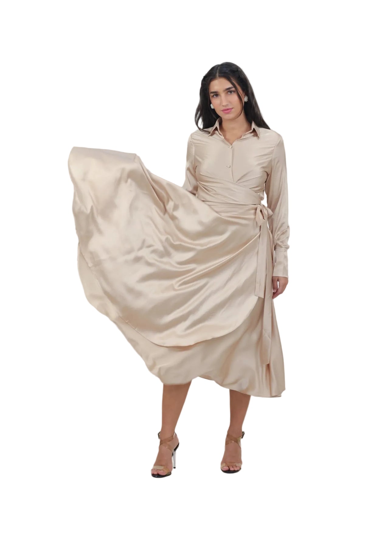 A woman modeling a long-sleeve beige satin midi dress with a wrap-style waist and tie belt, captured mid-motion as she swirls the fabric. The background is a solid, warm neutral beige studio backdrop.