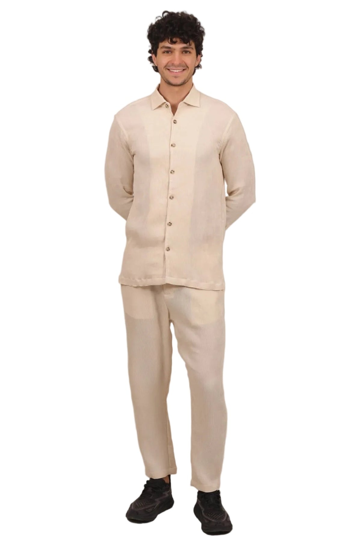 A man modeling a light beige two-piece linen set consisting of a long-sleeve button-up shirt and matching trousers, standing against a solid white background.