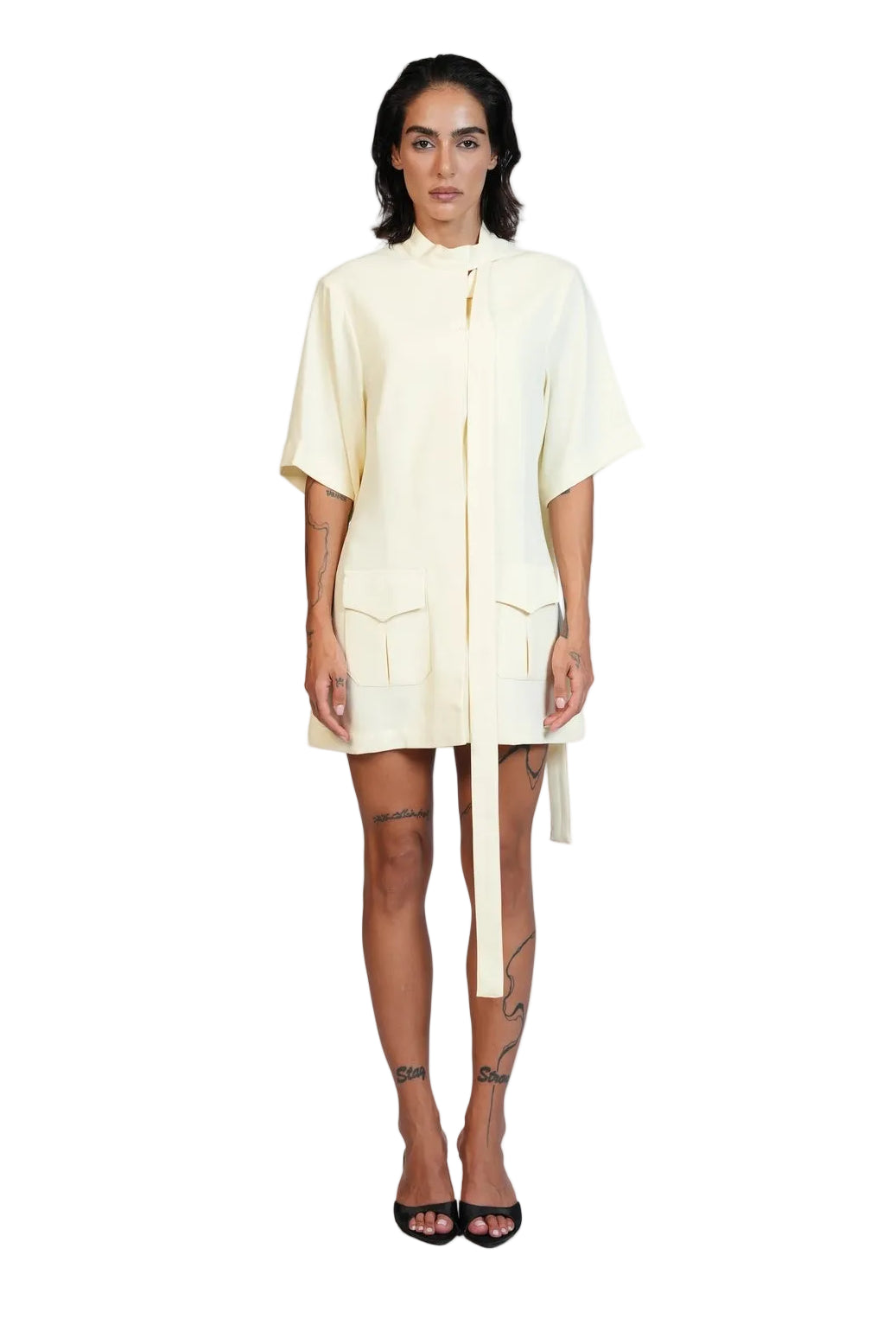 A model stands against a plain white studio background wearing a short-sleeved cream-colored shirt dress with a high collar, front zipper, and a long tie detail at the waist.