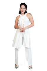 A woman modeling a white sleeveless halter top with a front keyhole cutout and flared tunic layer, paired with white wide-leg pants and silver pointed heels, against a plain white background.