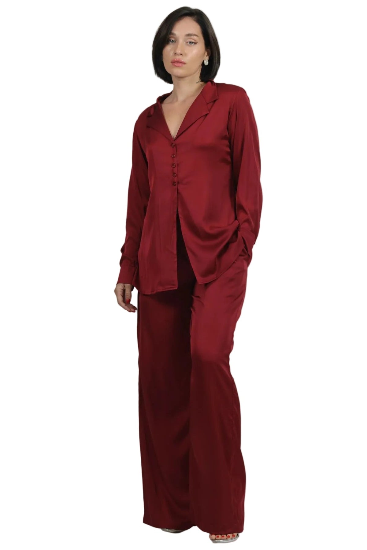 A woman modeling a matching two-piece satin pajama set in deep burgundy, consisting of a long-sleeve button-up top and wide-leg pants, shot against a solid white background.
