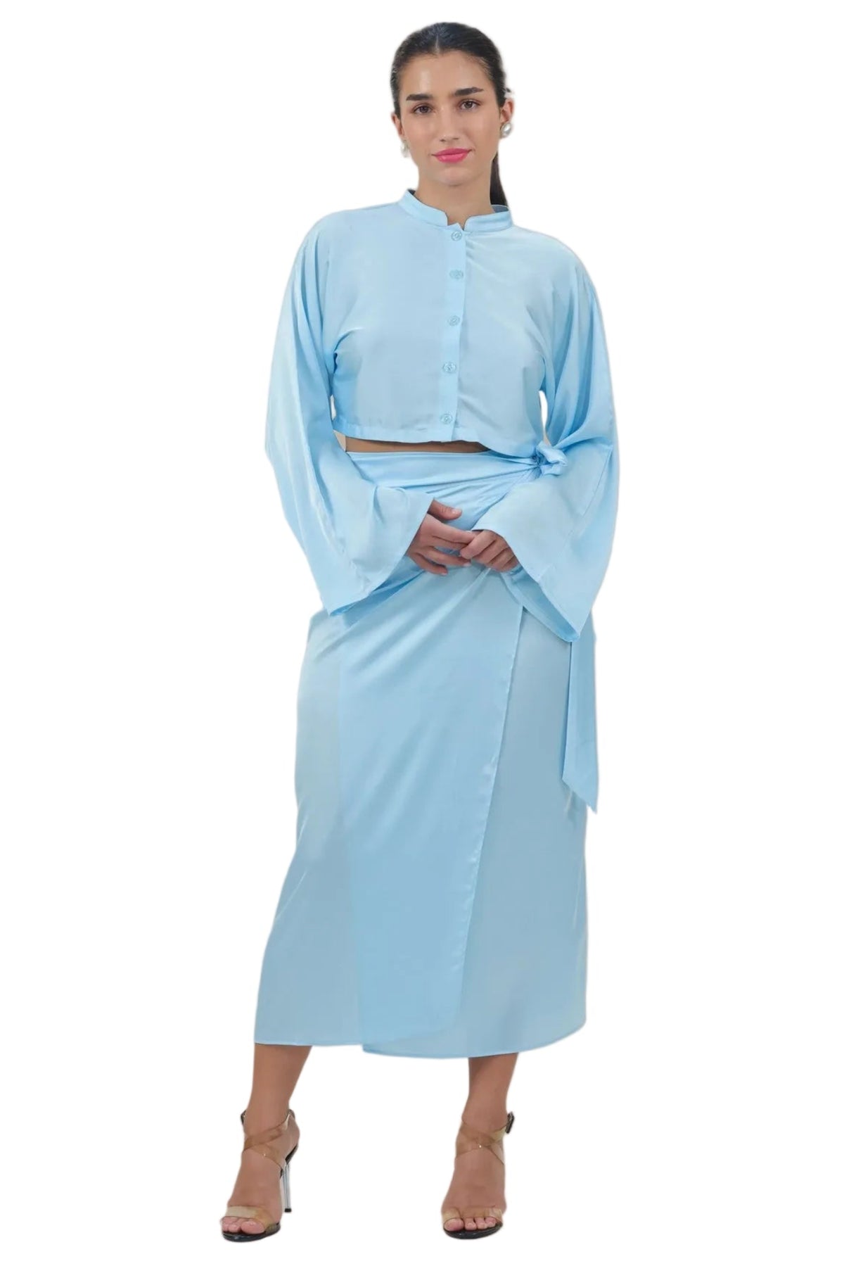 A woman modeling a light blue two-piece outfit consisting of a long-sleeve cropped button-front top and a matching midi wrap skirt, shown against a plain white background.