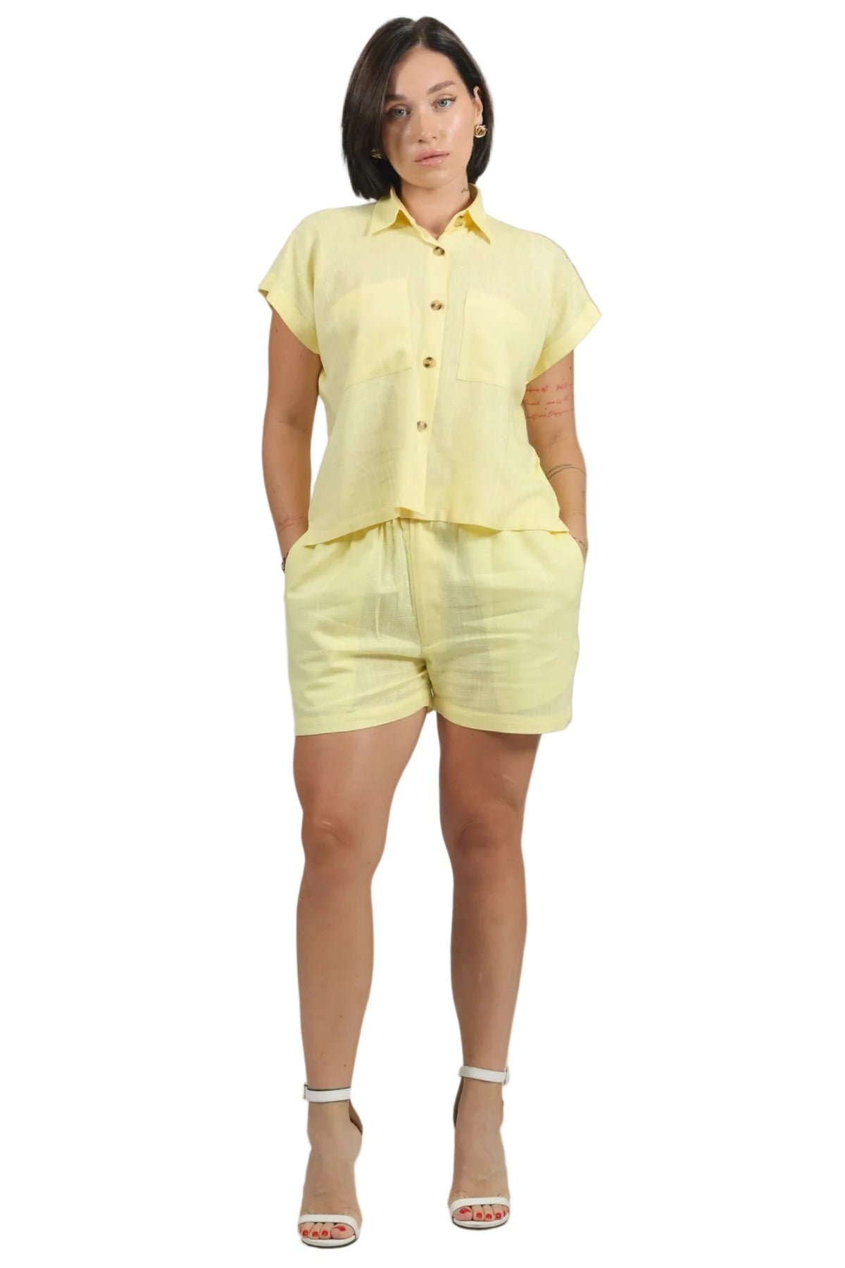 A woman modeling a light yellow short-sleeve button-up shirt and matching shorts set, paired with white heeled sandals, against a plain white background.