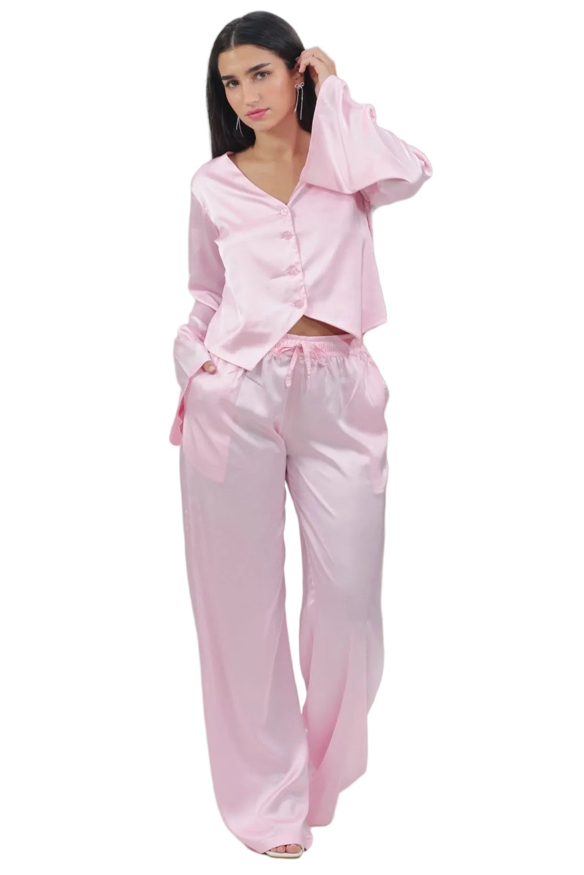 A woman modeling a light pink two-piece pajama set consisting of a long-sleeve button-up top and matching wide-leg pants, shot against a solid white background.