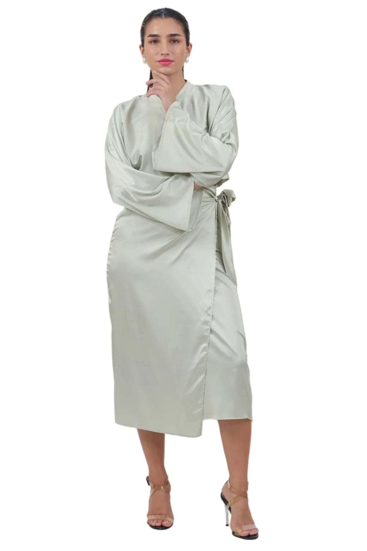 A woman modeling a light sage green wrap-style dress with long draped sleeves and a matching tie belt, photographed against a plain white studio background.
