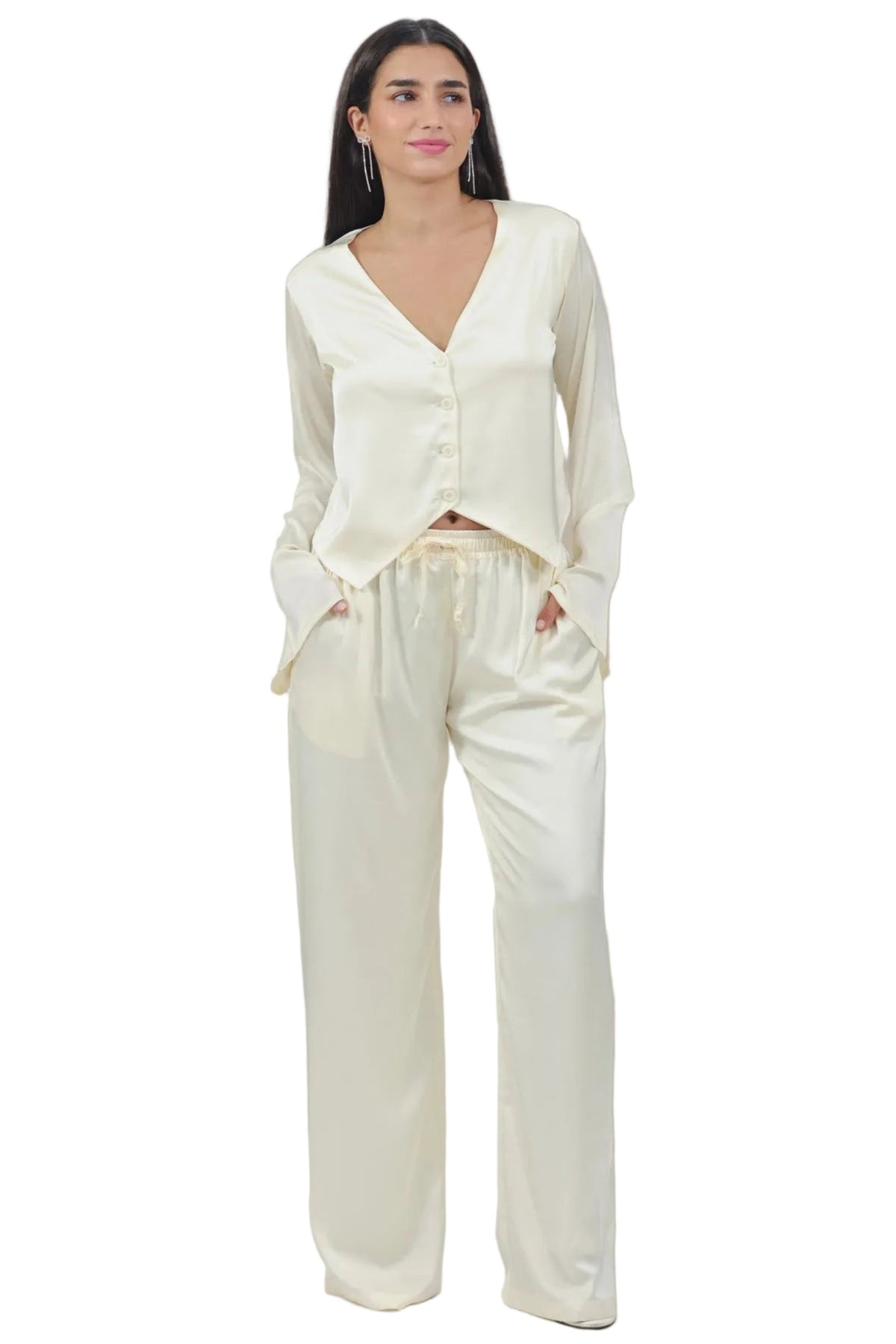 A woman modeling a cream-colored satin pajama set consisting of a long-sleeve button-up top and matching wide-leg pants, shot against a solid white background.