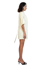 A woman in profile wearing a short cream-colored dress with a high collar and a long tie detail at the back, paired with black heeled sandals, against a plain white studio background.