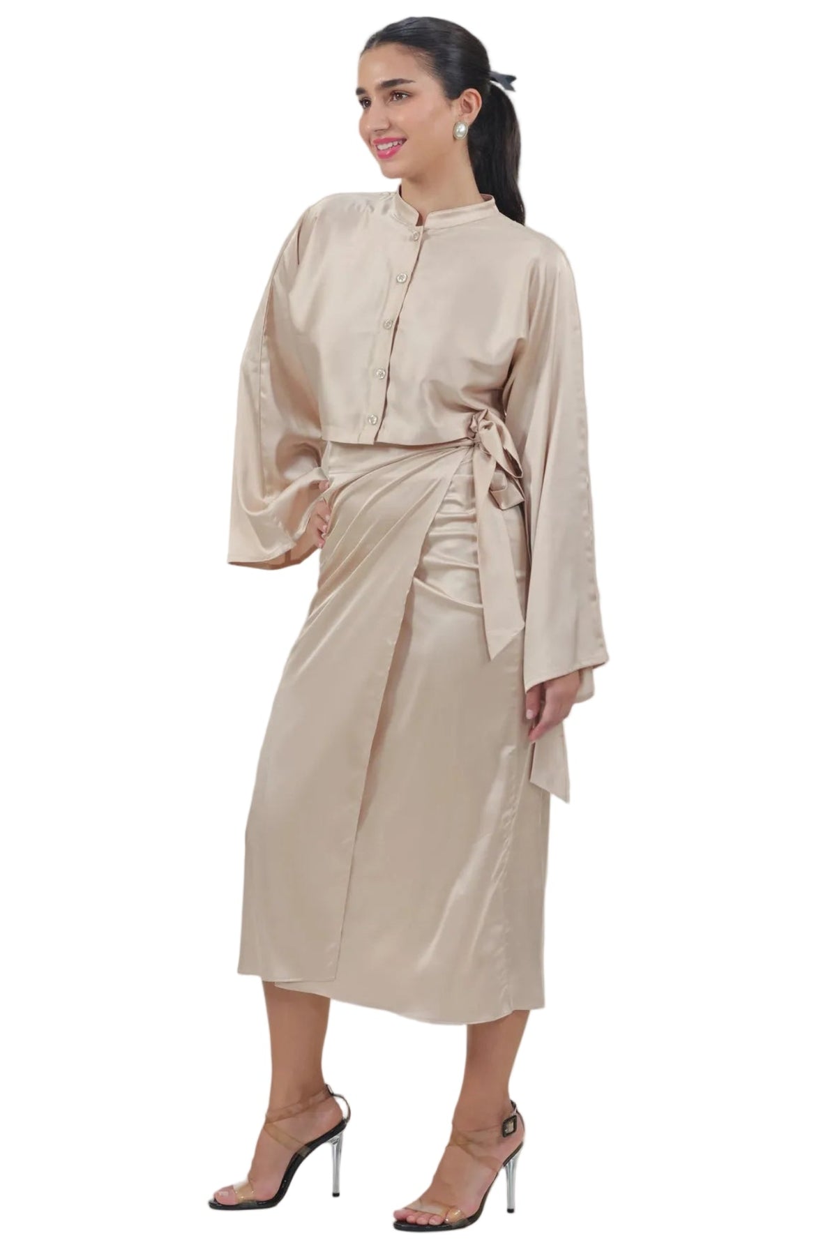 A woman modeling a beige satin-like long-sleeve tunic dress with a matching tie belt, paired with clear high-heeled sandals, against a plain white background.