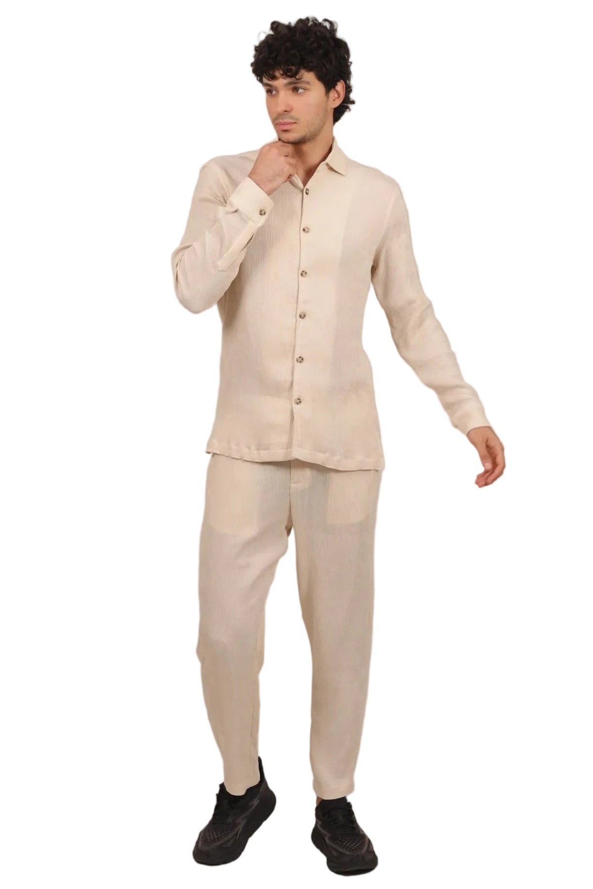 A man modeling a light beige two-piece linen set consisting of a long-sleeve button-up shirt and matching trousers, paired with black shoes, against a solid white background.